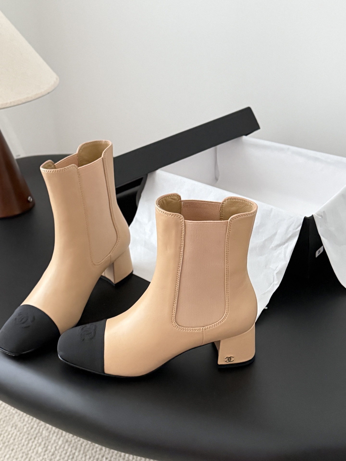 Chanel beige leather ankle boots with black toe caps and block heels displayed next to a black Chanel shoe box. - Hình 2