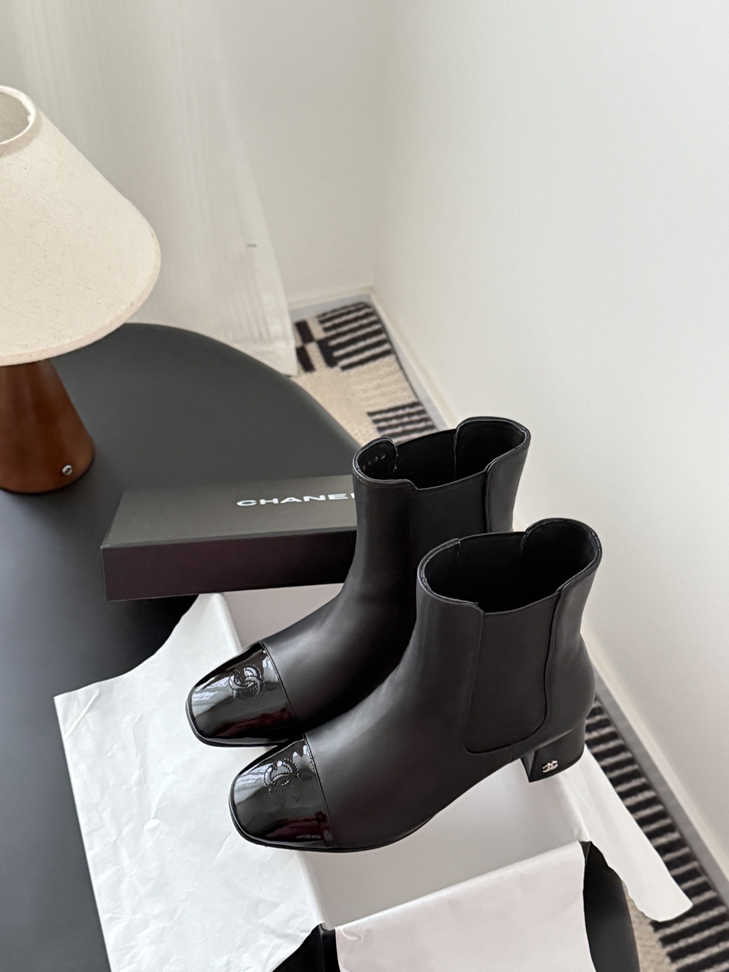 Pair of black Chanel leather ankle boots with patent leather cap toes and block heels displayed next to a Chanel shoe box. - Hình 9