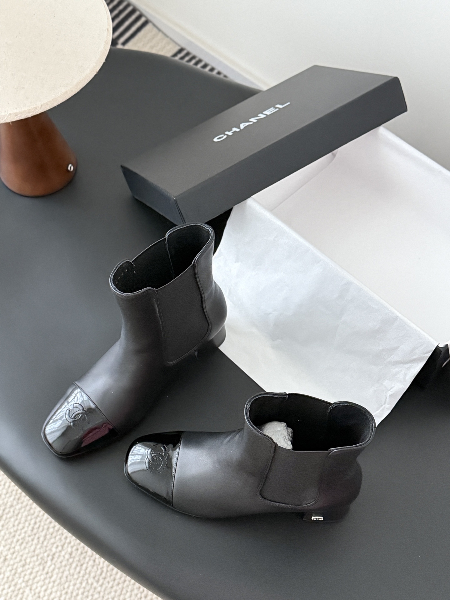 Pair of black Chanel leather ankle boots with patent leather cap toes and block heels displayed next to a Chanel shoe box. - Hình 8