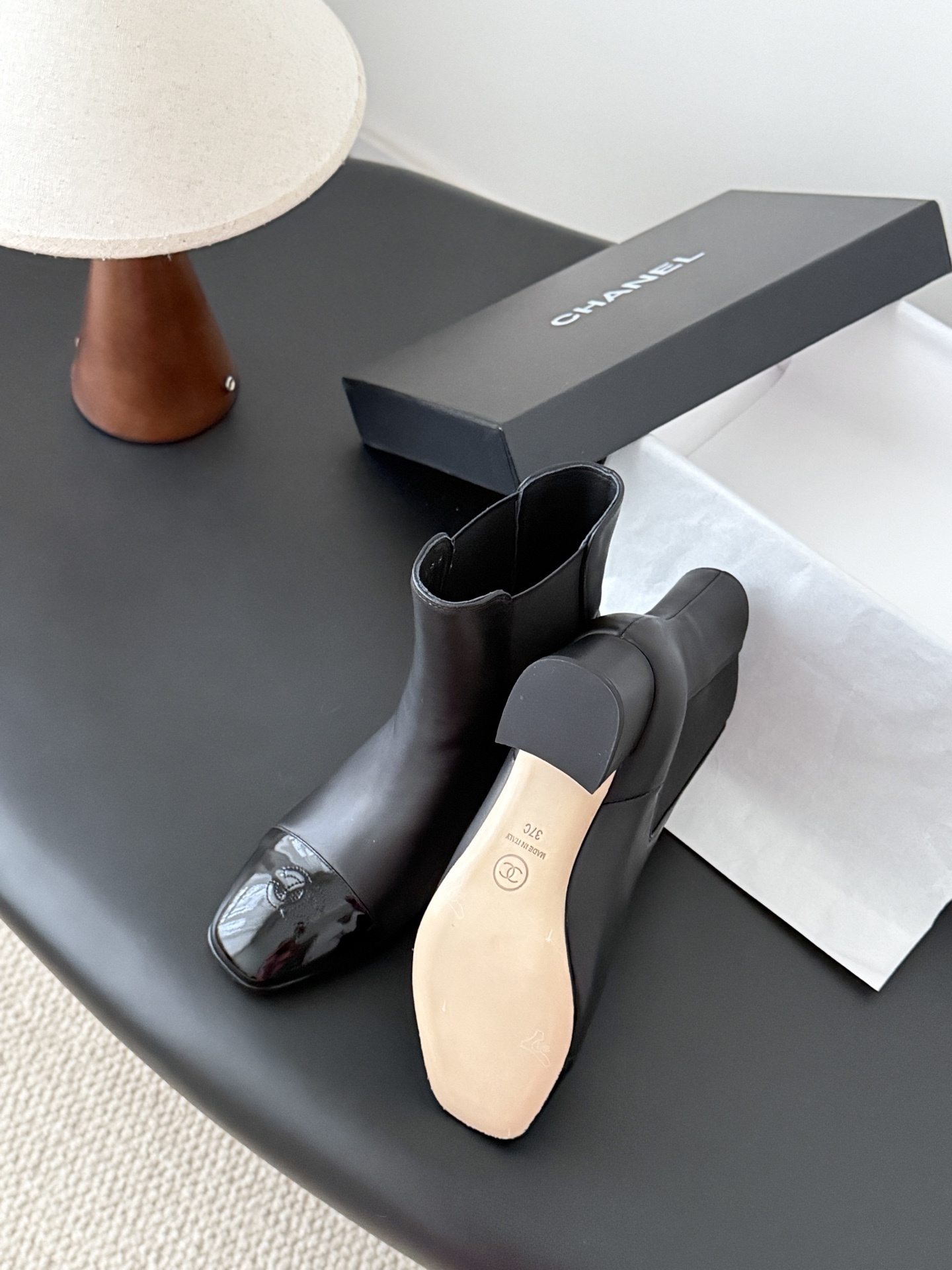 Pair of black Chanel leather ankle boots with patent leather cap toes and block heels displayed next to a Chanel shoe box. - Hình 6