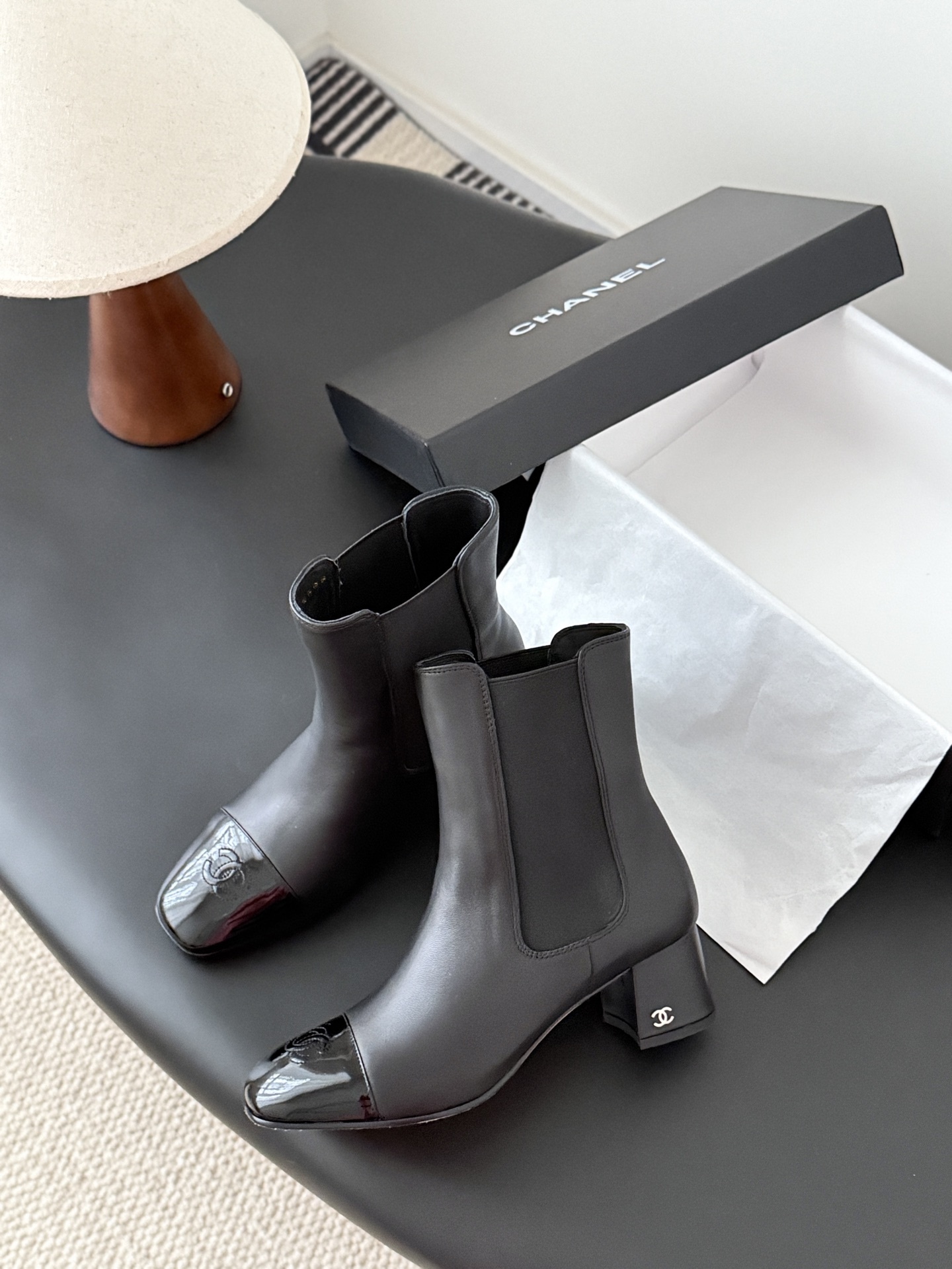 Pair of black Chanel leather ankle boots with patent leather cap toes and block heels displayed next to a Chanel shoe box. - Hình 4