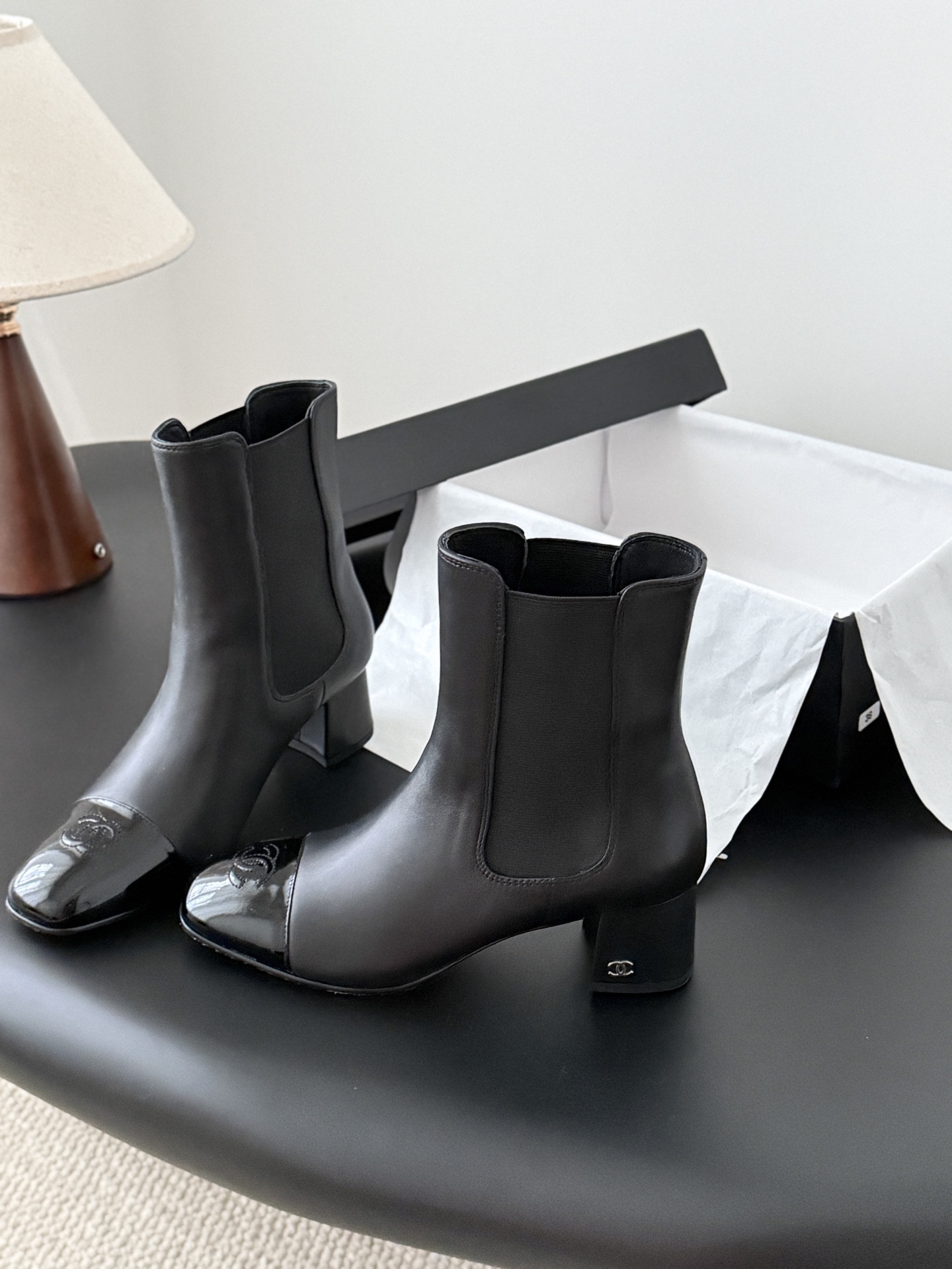 Pair of black Chanel leather ankle boots with patent leather cap toes and block heels displayed next to a Chanel shoe box. - Hình 2