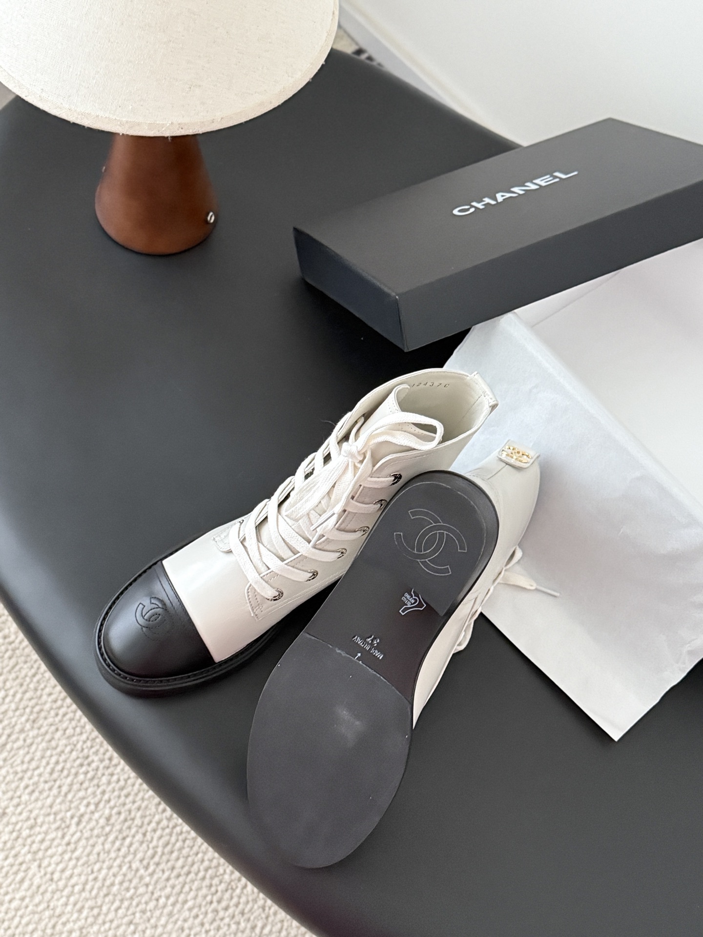 Chanel beige leather combat boots with black toe caps and gold CC logo on the heel next to a Chanel box. - Hình 5