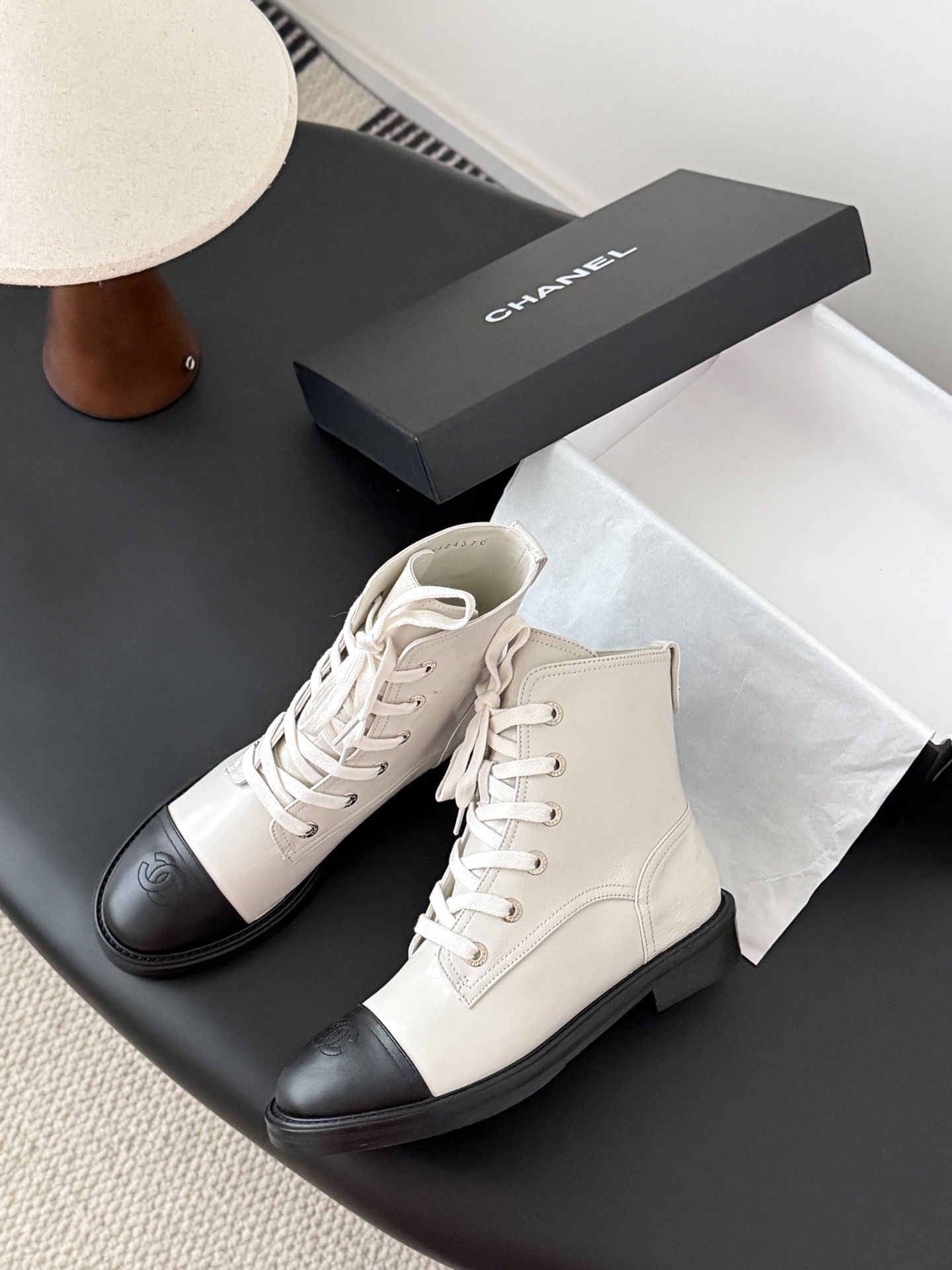 Chanel beige leather combat boots with black toe caps and gold CC logo on the heel next to a Chanel box. - Hình 4