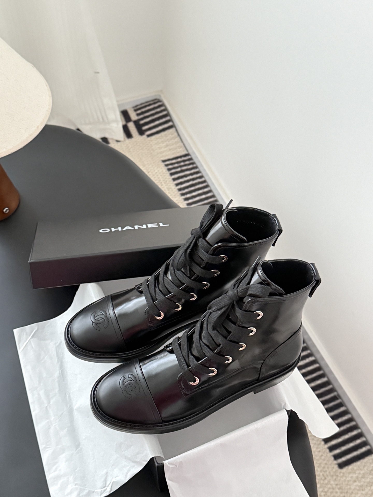 Pair of black Chanel patent leather lace-up combat boots with CC logo on toe and heel, displayed next to a Chanel shoebox on a black table. - Hình 9