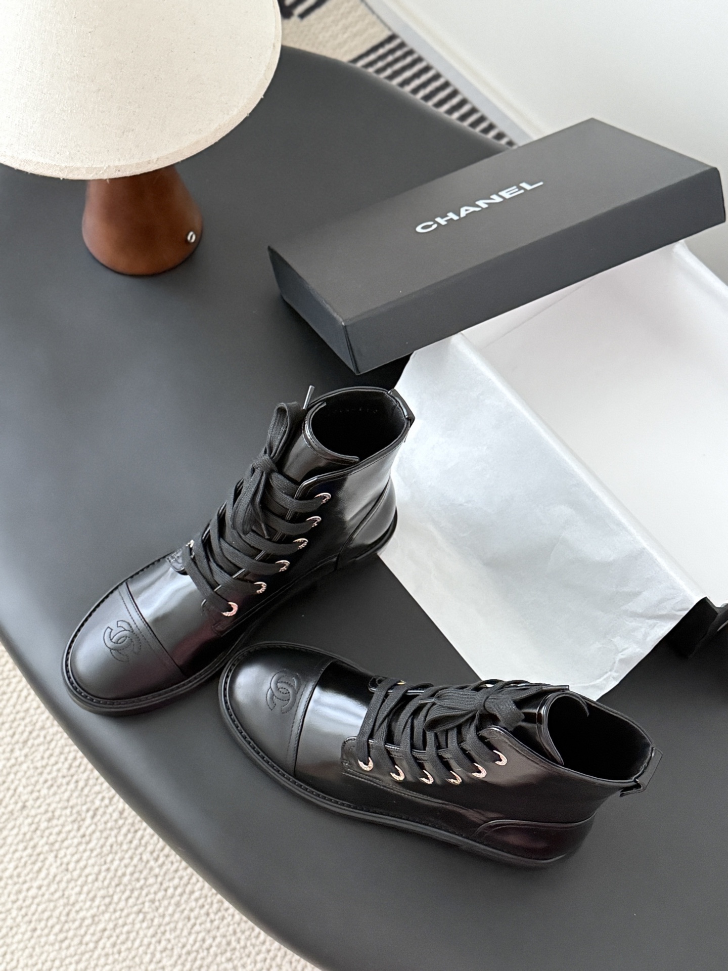 Pair of black Chanel patent leather lace-up combat boots with CC logo on toe and heel, displayed next to a Chanel shoebox on a black table. - Hình 6