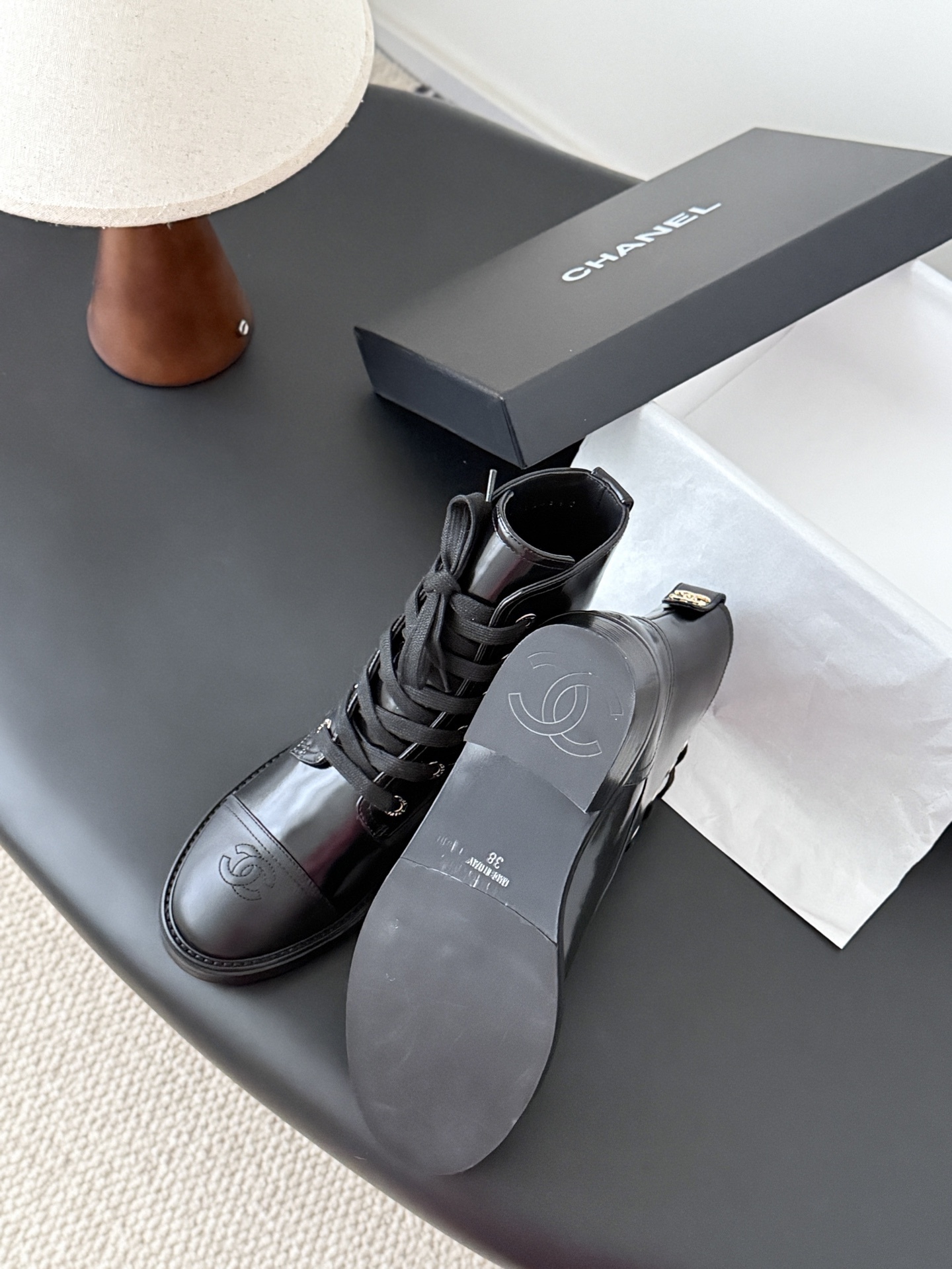 Pair of black Chanel patent leather lace-up combat boots with CC logo on toe and heel, displayed next to a Chanel shoebox on a black table. - Hình 5