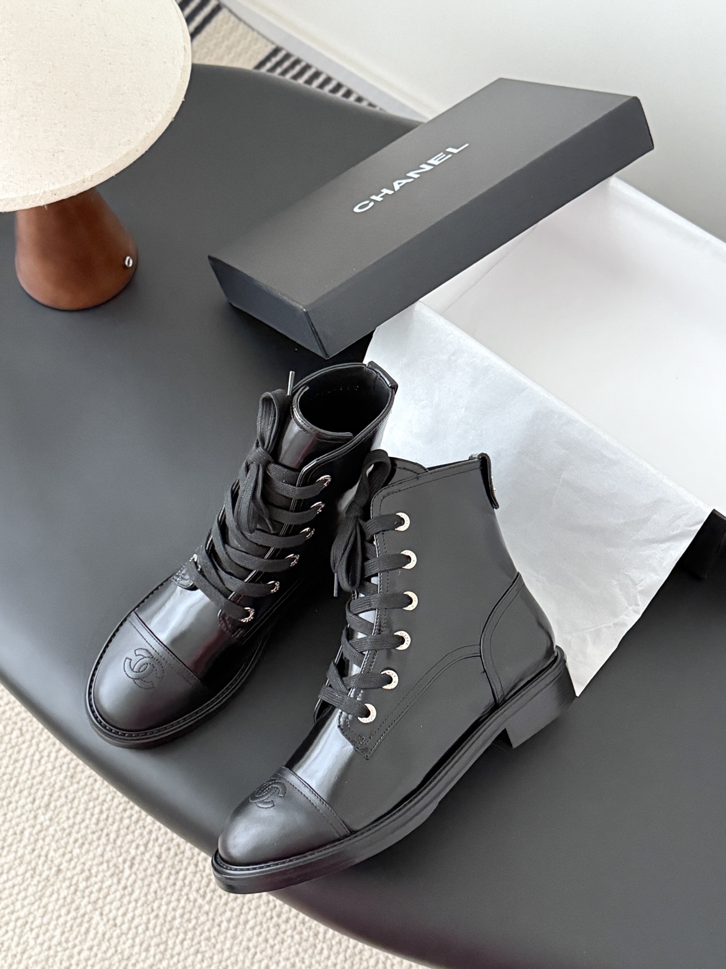 Pair of black Chanel patent leather lace-up combat boots with CC logo on toe and heel, displayed next to a Chanel shoebox on a black table. - Hình 4