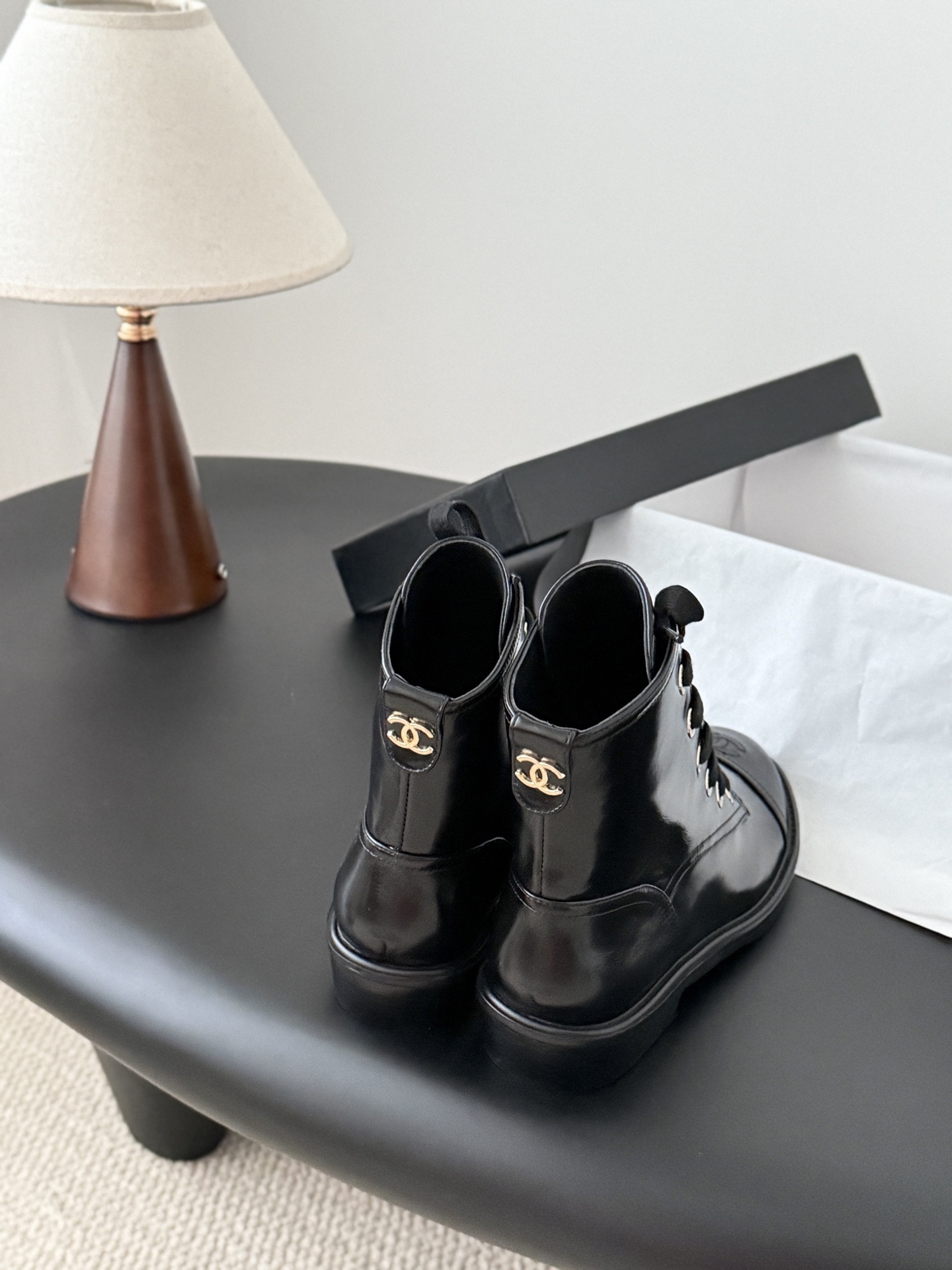 Pair of black Chanel patent leather lace-up combat boots with CC logo on toe and heel, displayed next to a Chanel shoebox on a black table. - Hình 3