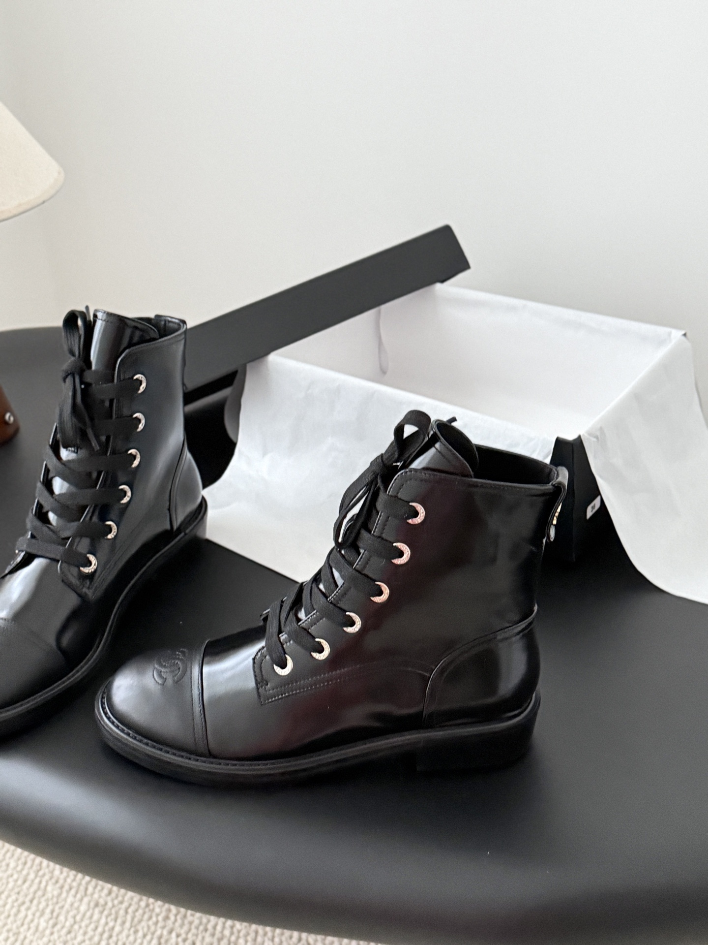 Pair of black Chanel patent leather lace-up combat boots with CC logo on toe and heel, displayed next to a Chanel shoebox on a black table. - Hình 2