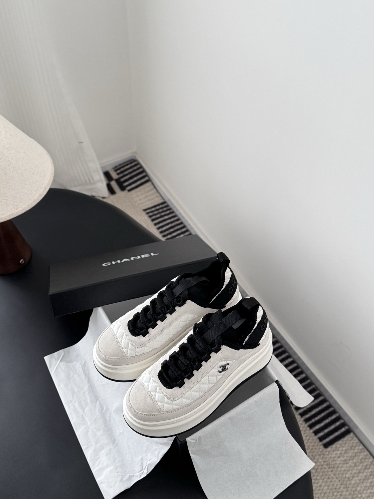 A pair of Chanel white and black quilted platform sneakers next to their original black Chanel box on a table. - Hình 6
