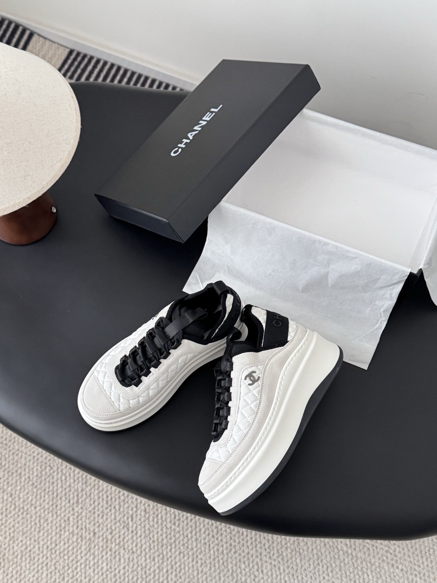 A pair of Chanel white and black quilted platform sneakers next to their original black Chanel box on a table. - Hình 3