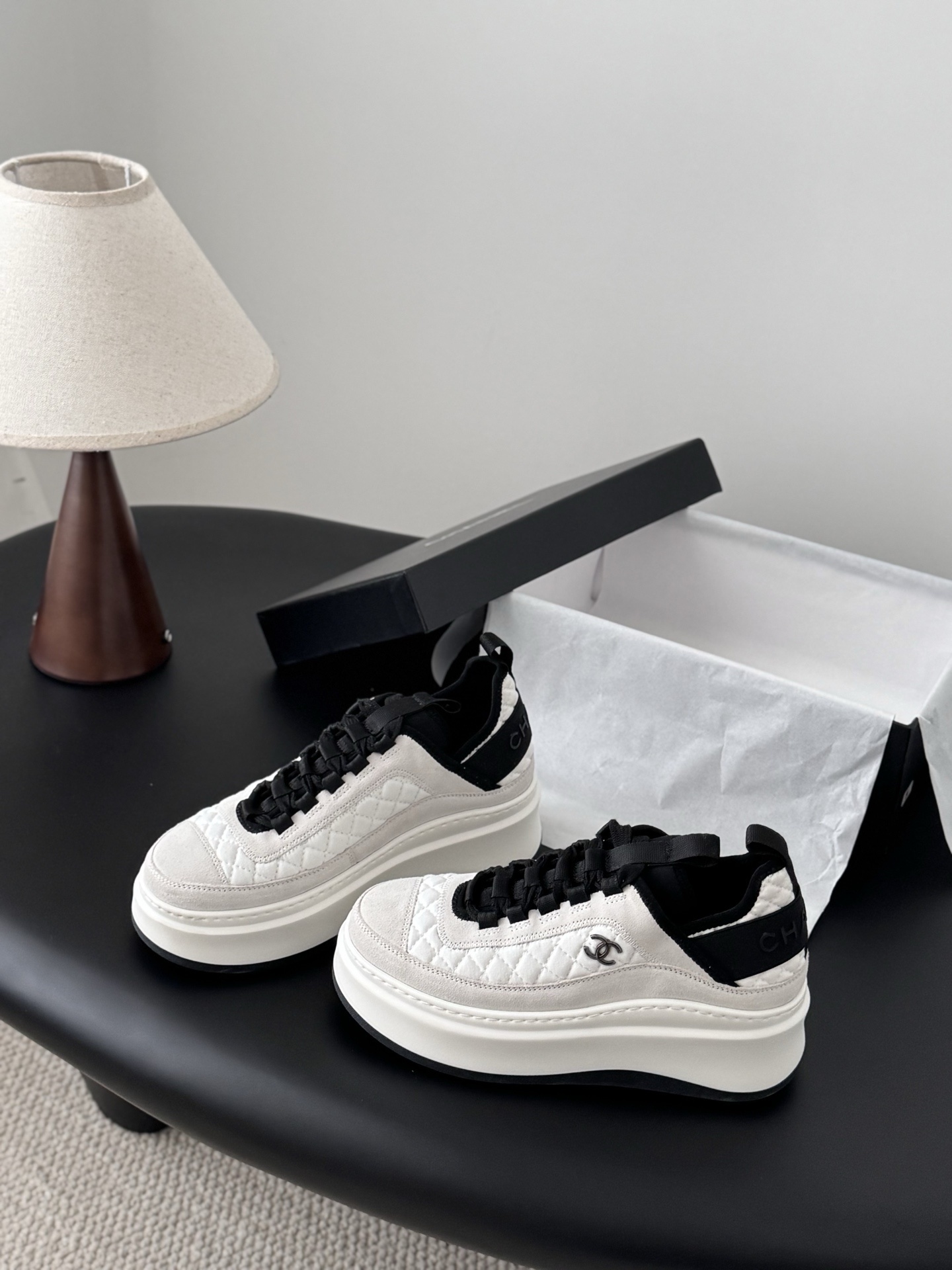 A pair of Chanel white and black quilted platform sneakers next to their original black Chanel box on a table. - Hình 2