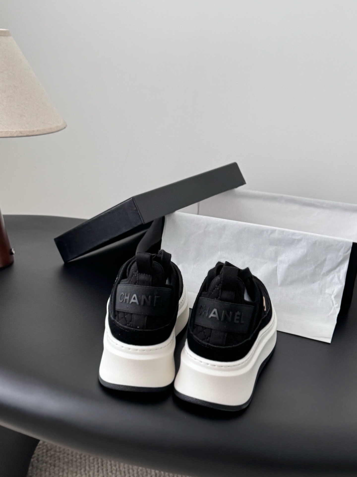 A pair of black Chanel quilted platform sneakers with white soles and gold CC logo next to a black Chanel shoe box. - Hình 5
