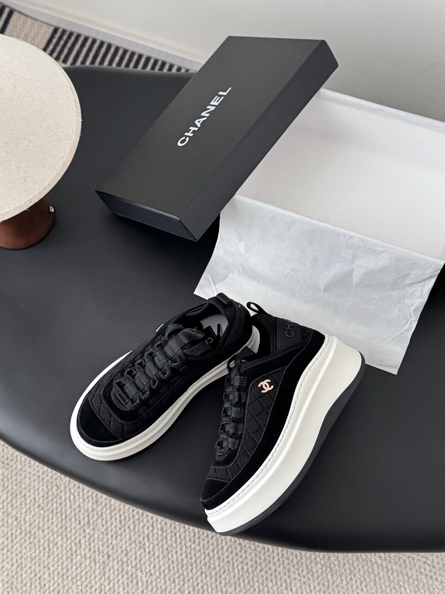 A pair of black Chanel quilted platform sneakers with white soles and gold CC logo next to a black Chanel shoe box. - Hình 3