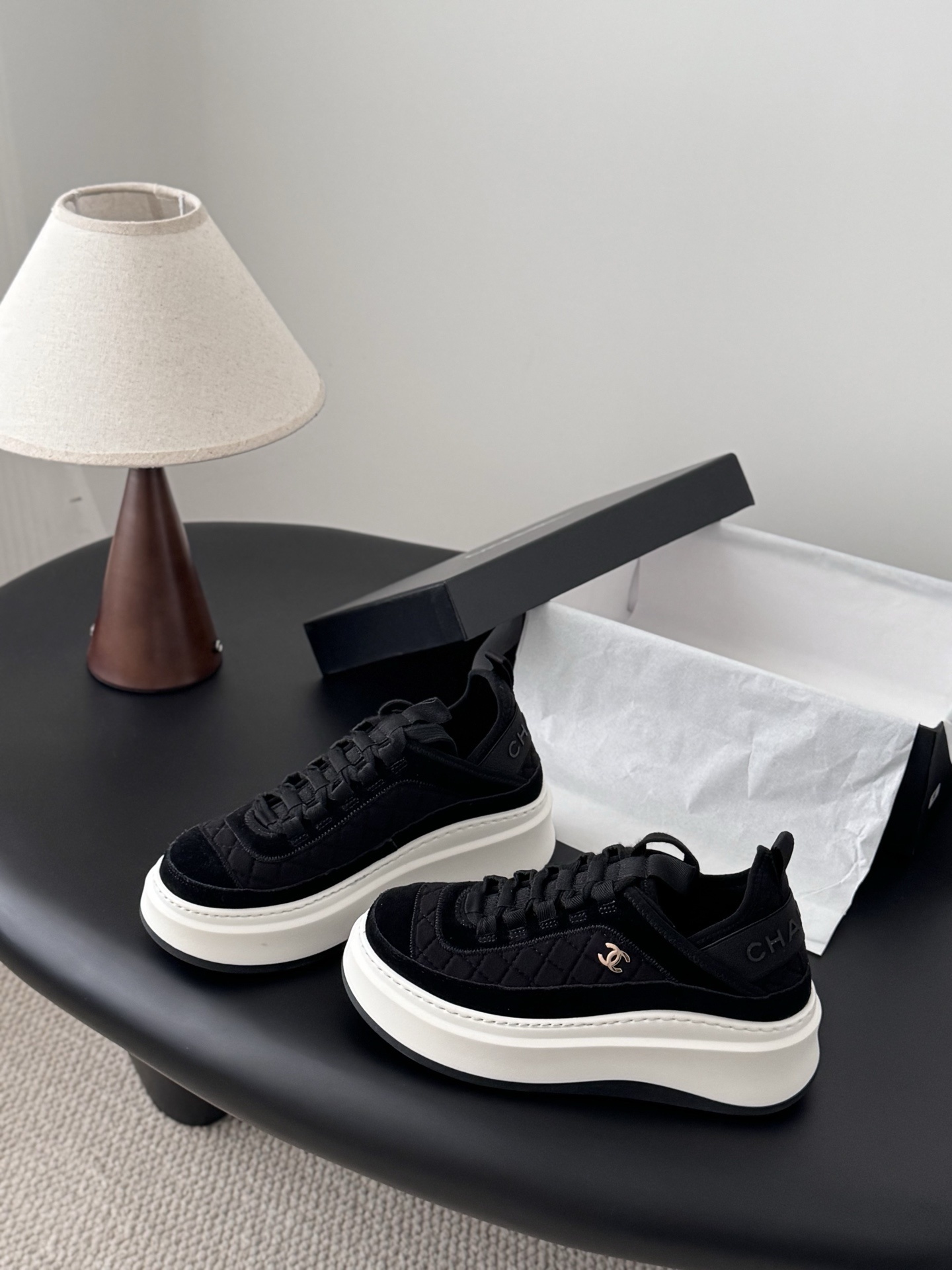 A pair of black Chanel quilted platform sneakers with white soles and gold CC logo next to a black Chanel shoe box. - Hình 2
