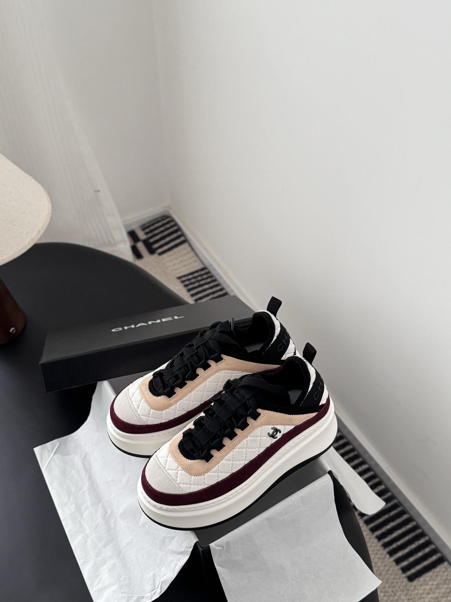 A pair of Chanel white quilted leather platform sneakers with burgundy and beige suede accents sitting next to a black Chanel shoe box. - Hình 6