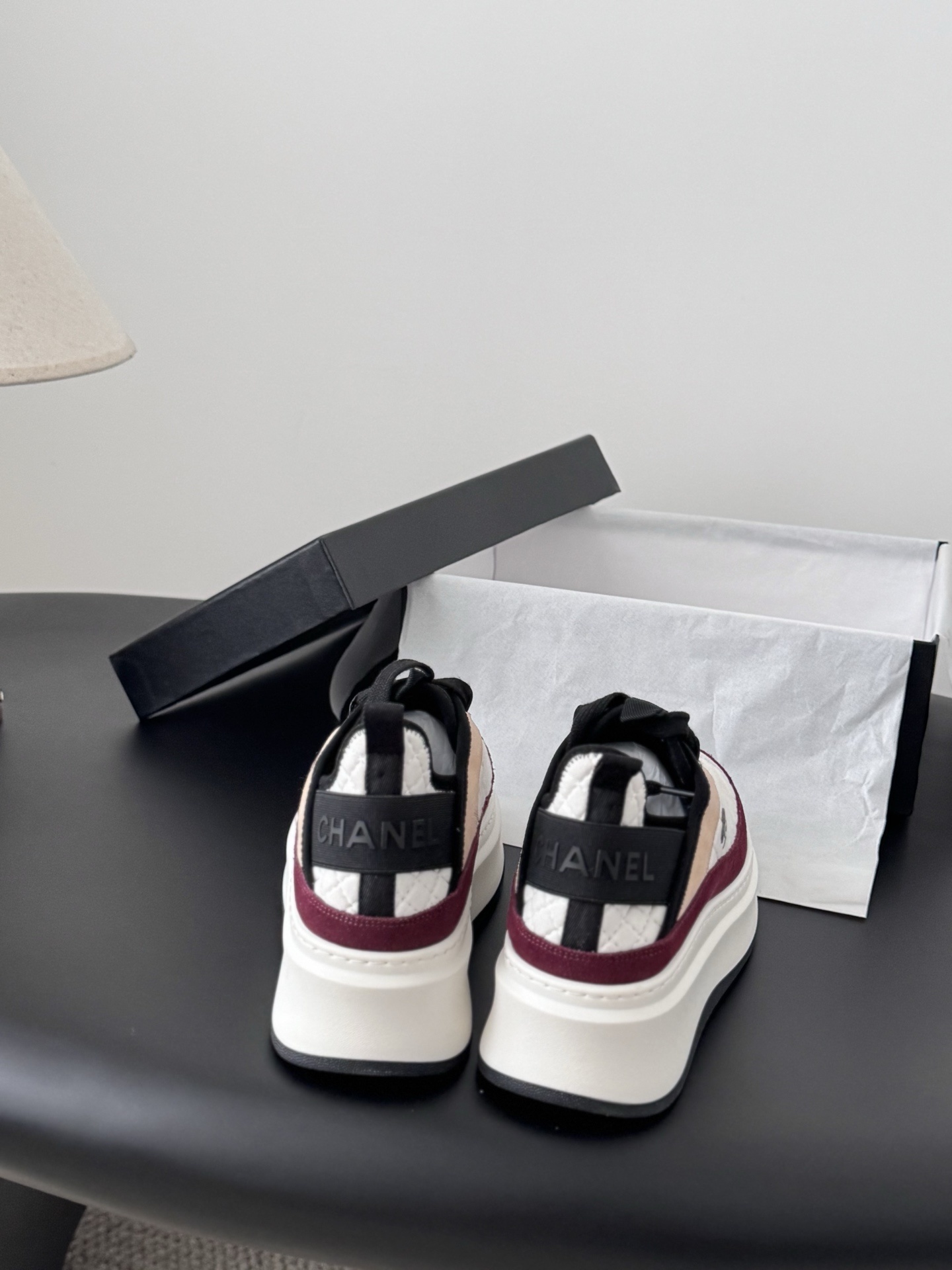 A pair of Chanel white quilted leather platform sneakers with burgundy and beige suede accents sitting next to a black Chanel shoe box. - Hình 5