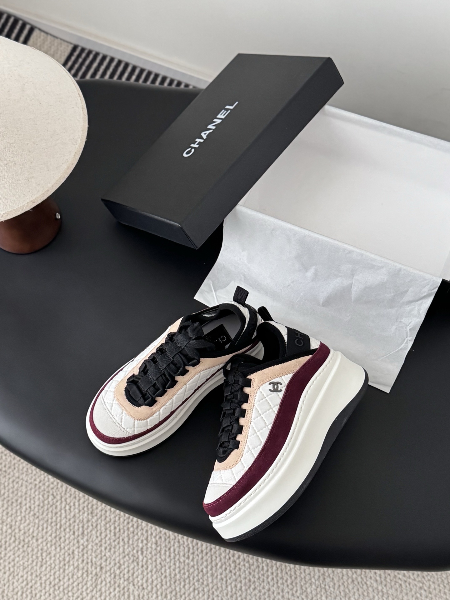 A pair of Chanel white quilted leather platform sneakers with burgundy and beige suede accents sitting next to a black Chanel shoe box. - Hình 3