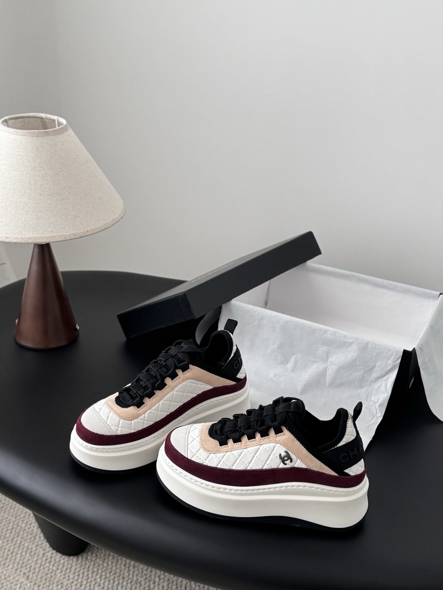 A pair of Chanel white quilted leather platform sneakers with burgundy and beige suede accents sitting next to a black Chanel shoe box. - Hình 2