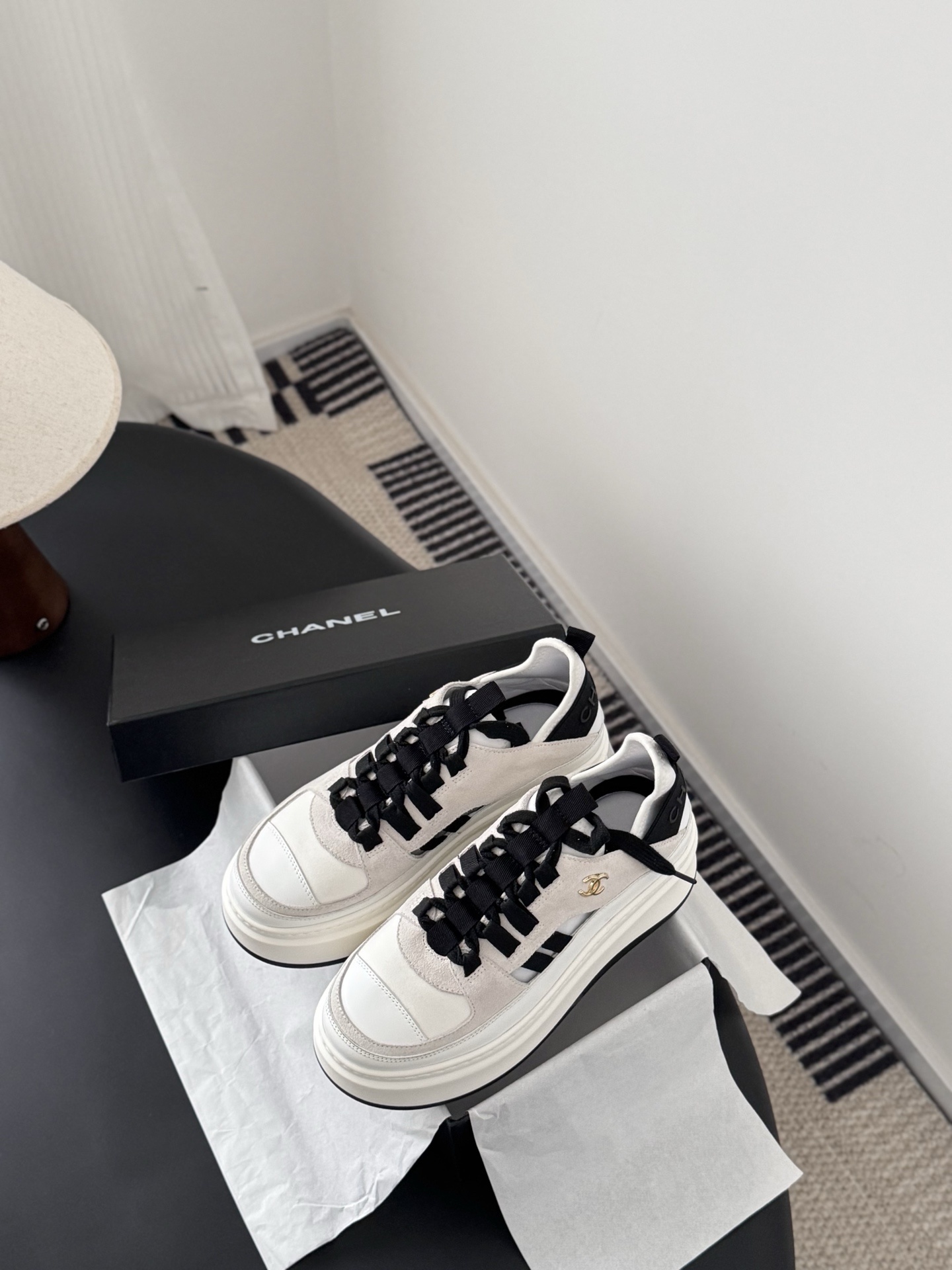 A pair of white and black Chanel platform sneakers with gold CC logo next to a black Chanel shoe box on a black table. - Hình 6