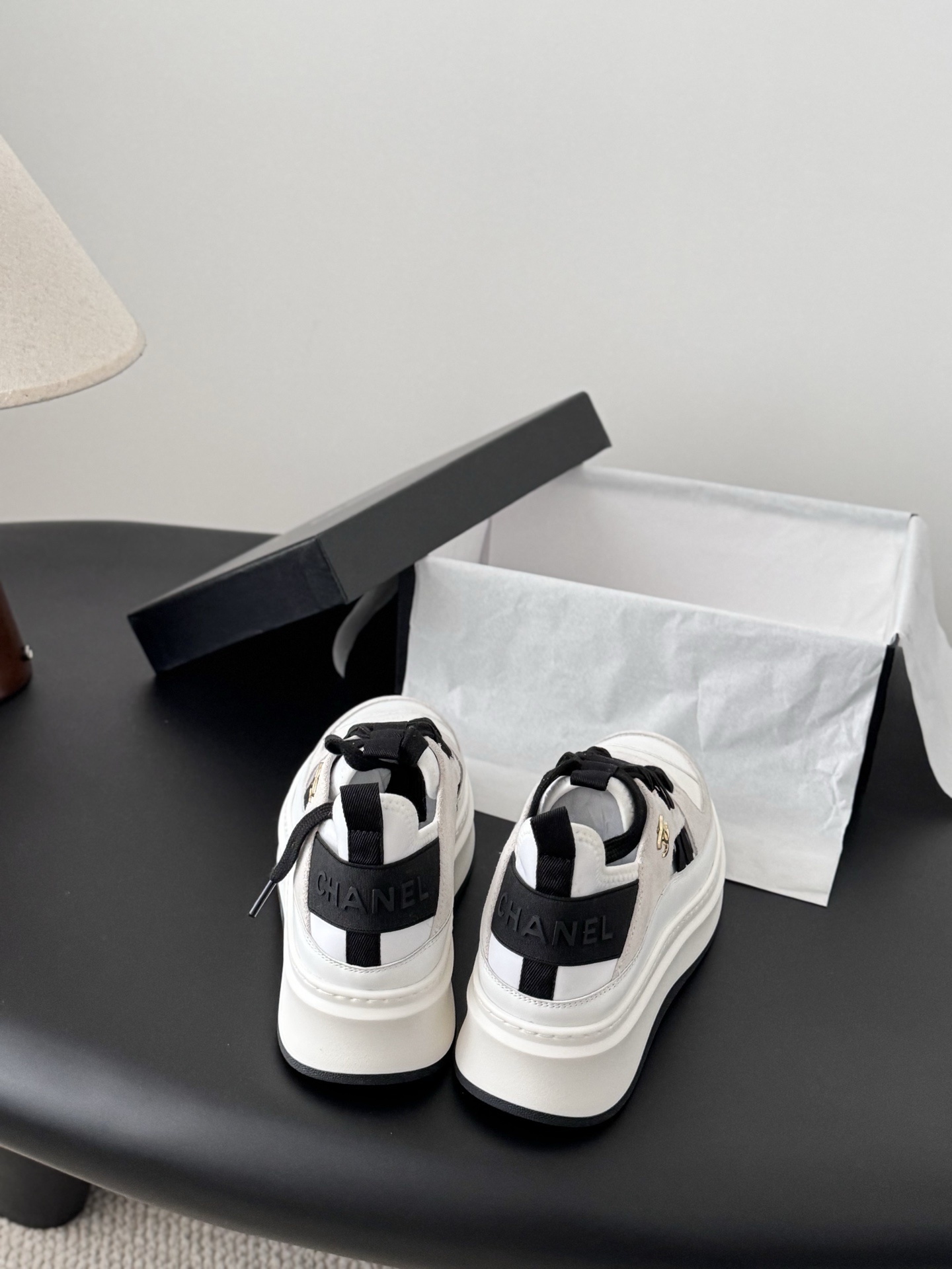 A pair of white and black Chanel platform sneakers with gold CC logo next to a black Chanel shoe box on a black table. - Hình 5
