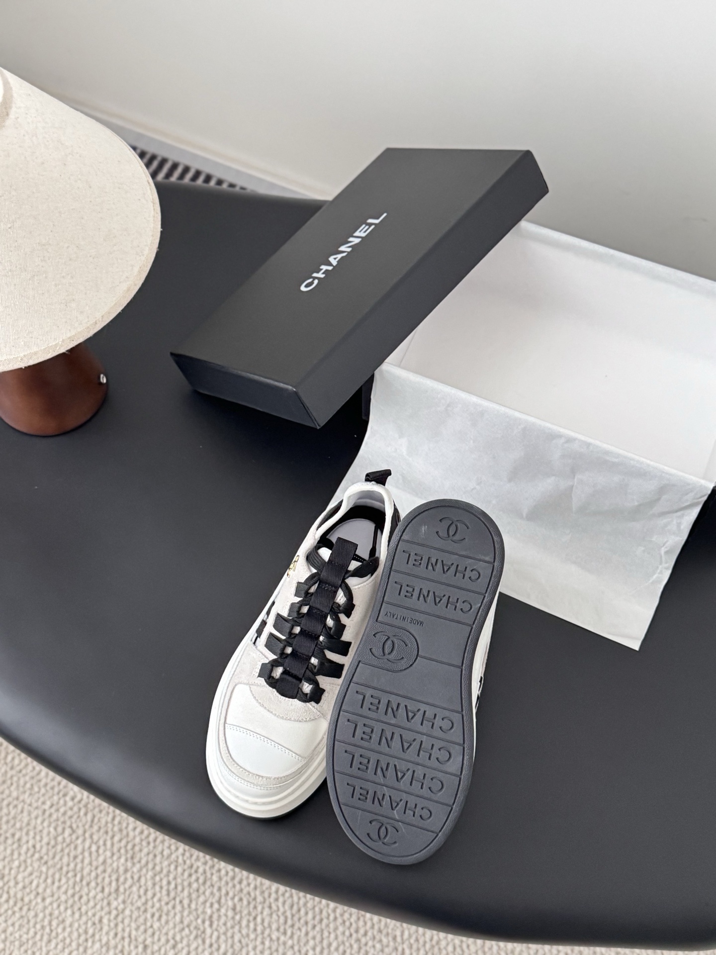 A pair of white and black Chanel platform sneakers with gold CC logo next to a black Chanel shoe box on a black table. - Hình 4