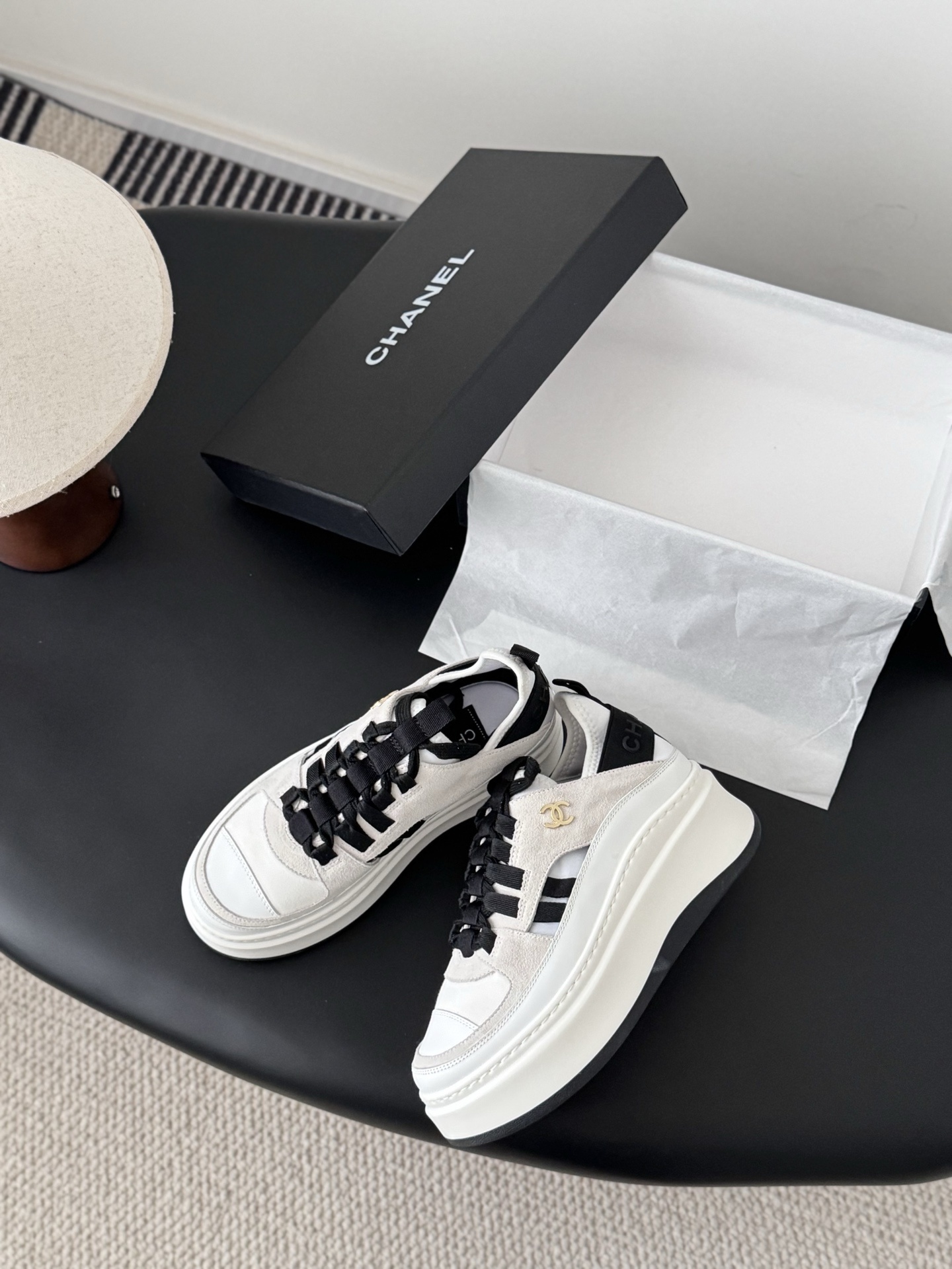 A pair of white and black Chanel platform sneakers with gold CC logo next to a black Chanel shoe box on a black table. - Hình 3