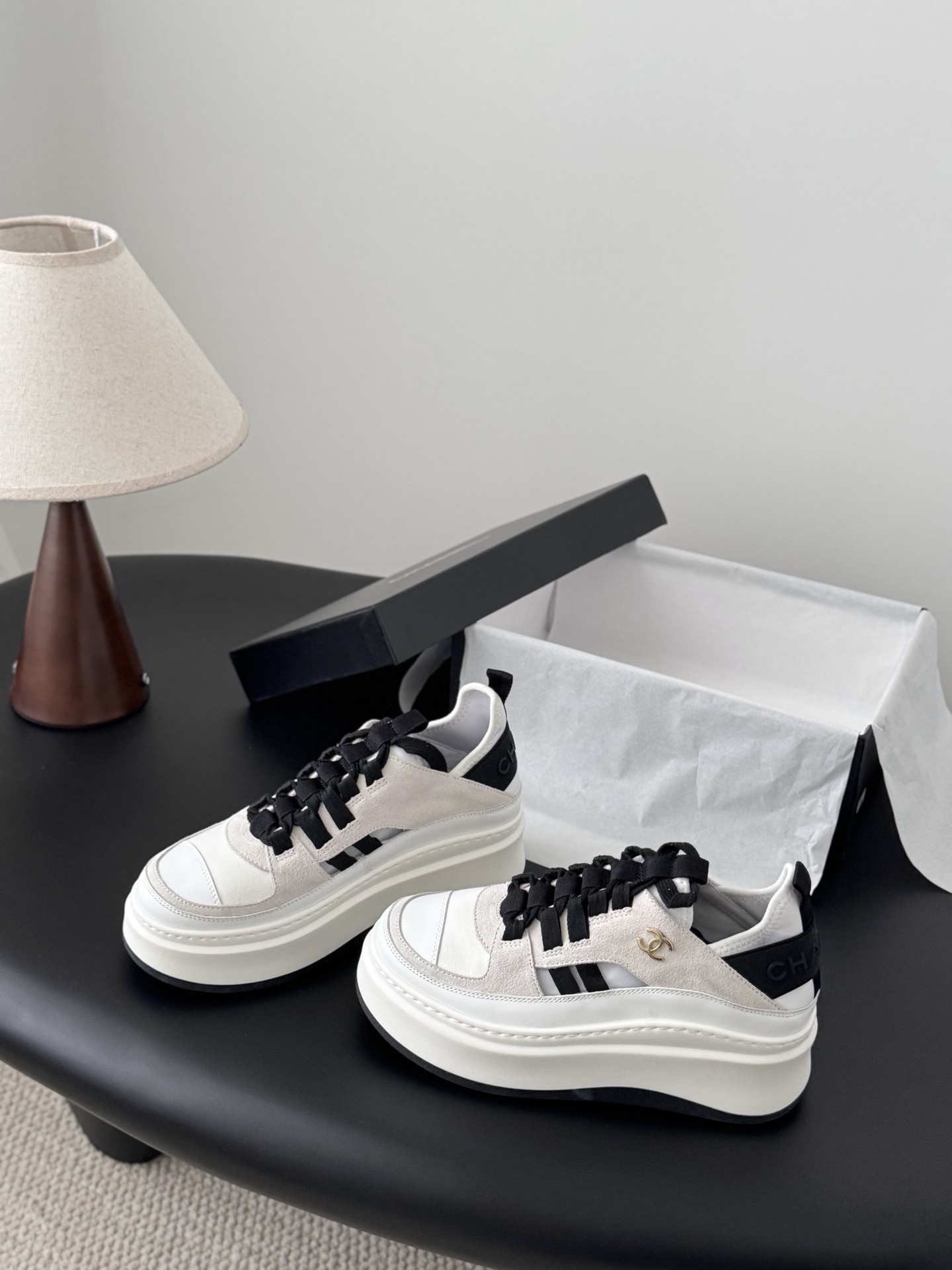 A pair of white and black Chanel platform sneakers with gold CC logo next to a black Chanel shoe box on a black table. - Hình 2