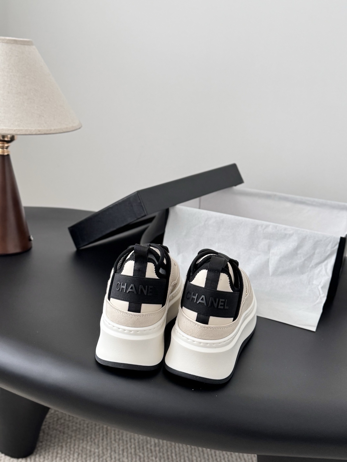 A pair of luxury Chanel beige and black platform sneakers with gold CC logo on a black surface next to a Chanel shoe box. - Hình 5