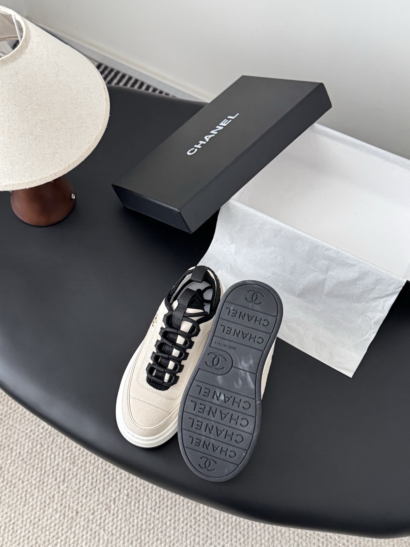 A pair of luxury Chanel beige and black platform sneakers with gold CC logo on a black surface next to a Chanel shoe box. - Hình 4