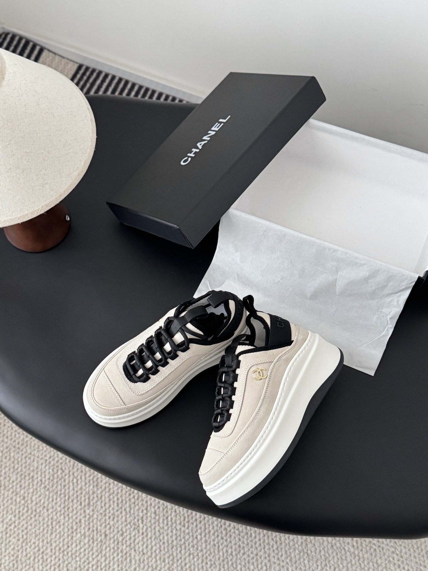 A pair of luxury Chanel beige and black platform sneakers with gold CC logo on a black surface next to a Chanel shoe box. - Hình 3