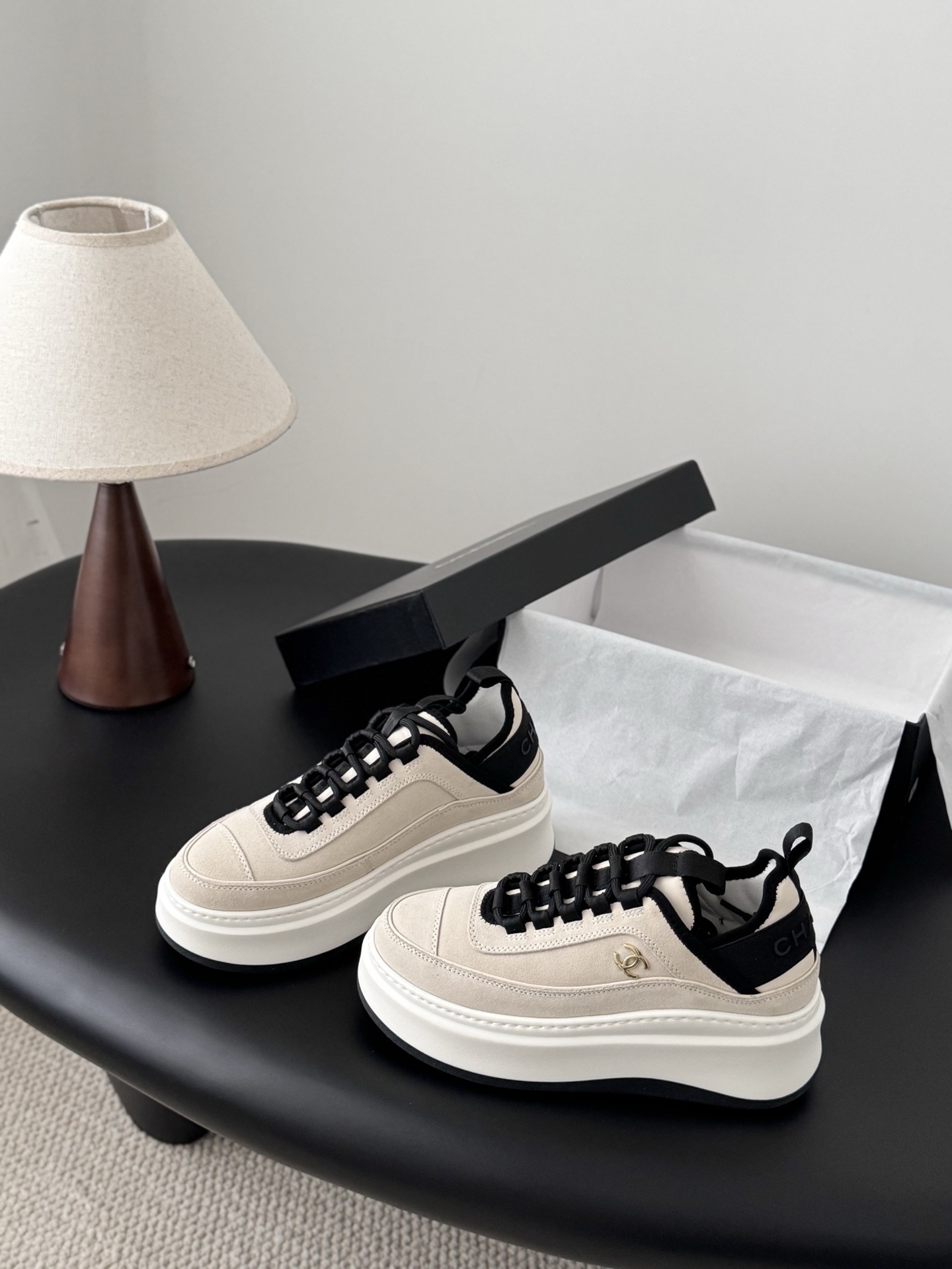 A pair of luxury Chanel beige and black platform sneakers with gold CC logo on a black surface next to a Chanel shoe box. - Hình 2