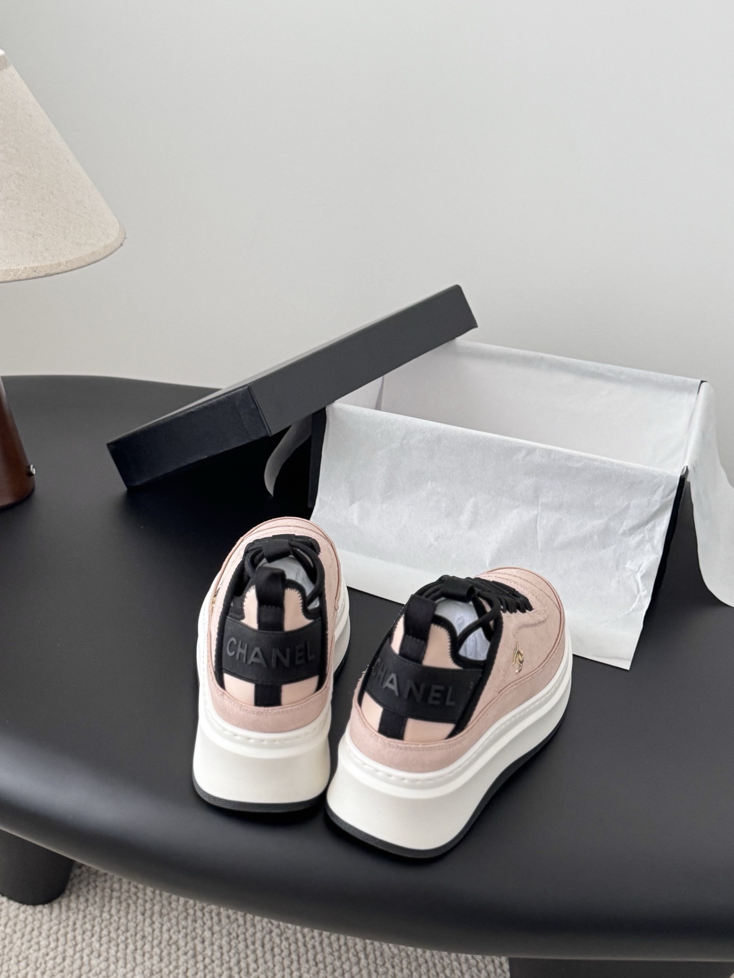 A pair of Chanel pink suede platform sneakers with black laces and gold CC logo on a black table next to a Chanel shoebox. - Hình 5