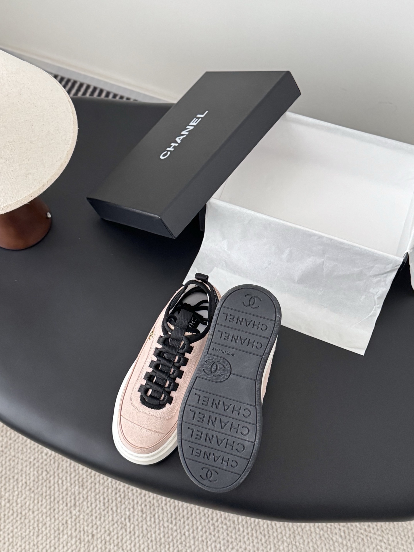 A pair of Chanel pink suede platform sneakers with black laces and gold CC logo on a black table next to a Chanel shoebox. - Hình 4