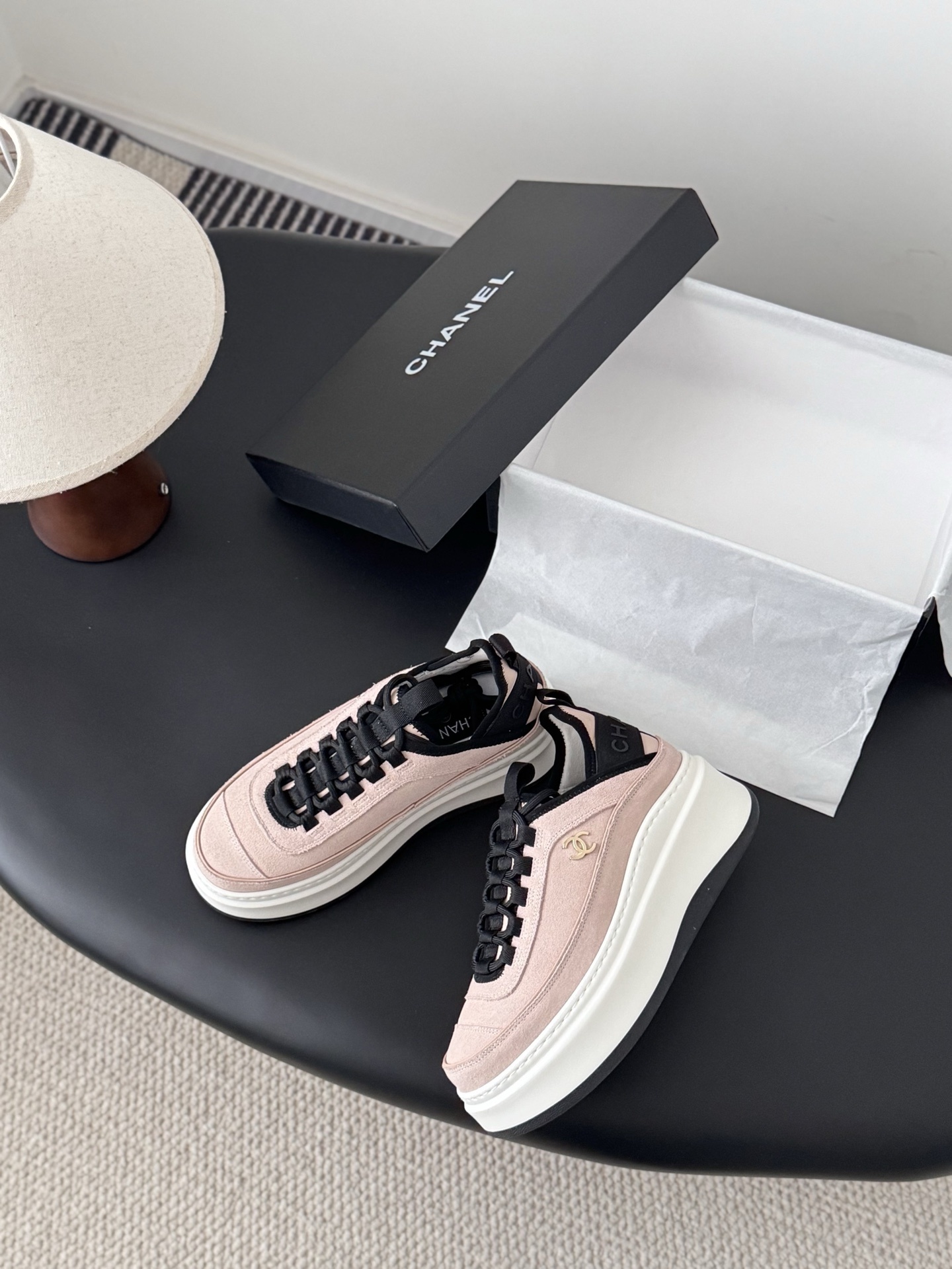 A pair of Chanel pink suede platform sneakers with black laces and gold CC logo on a black table next to a Chanel shoebox. - Hình 3