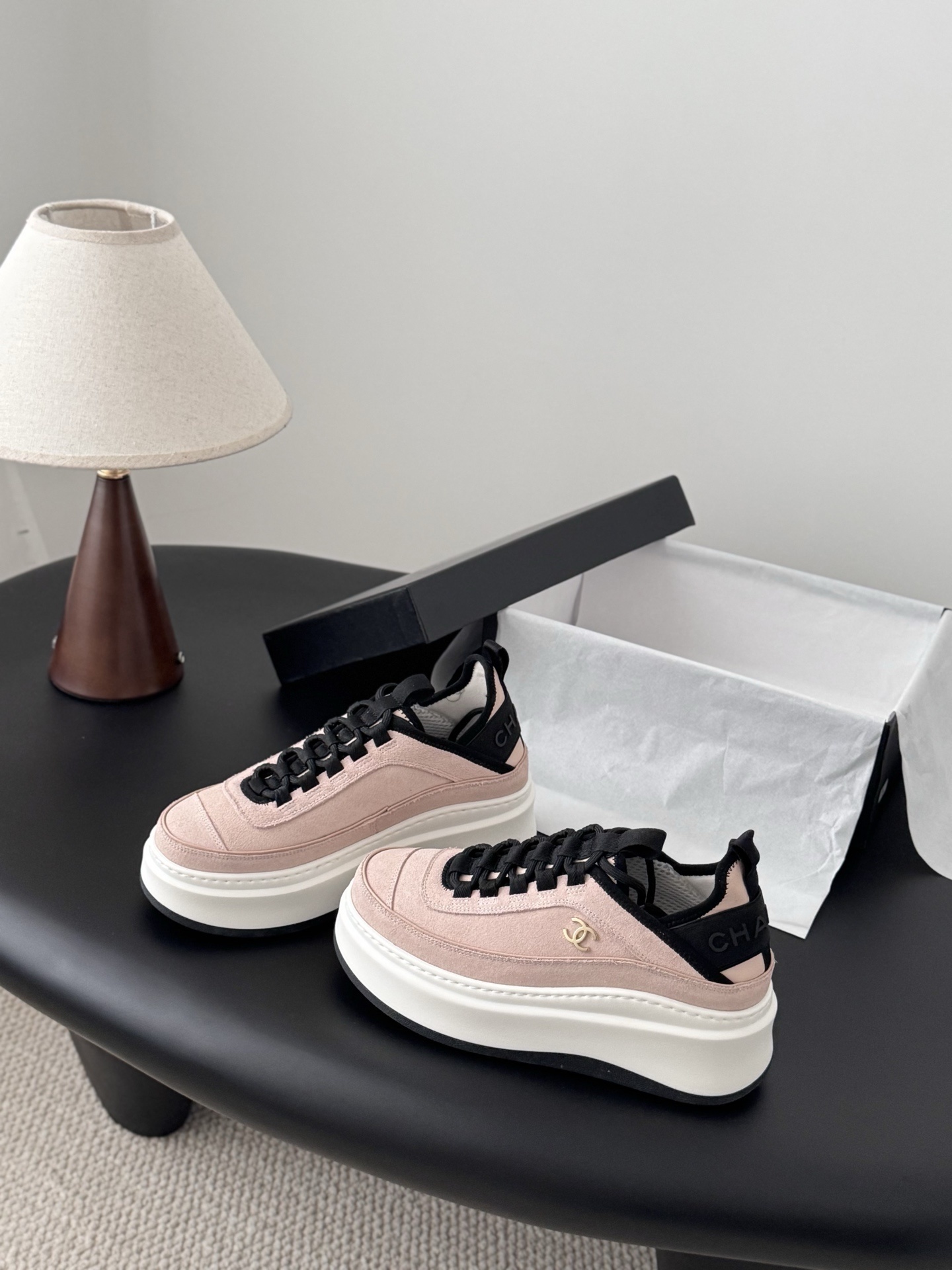 A pair of Chanel pink suede platform sneakers with black laces and gold CC logo on a black table next to a Chanel shoebox. - Hình 2