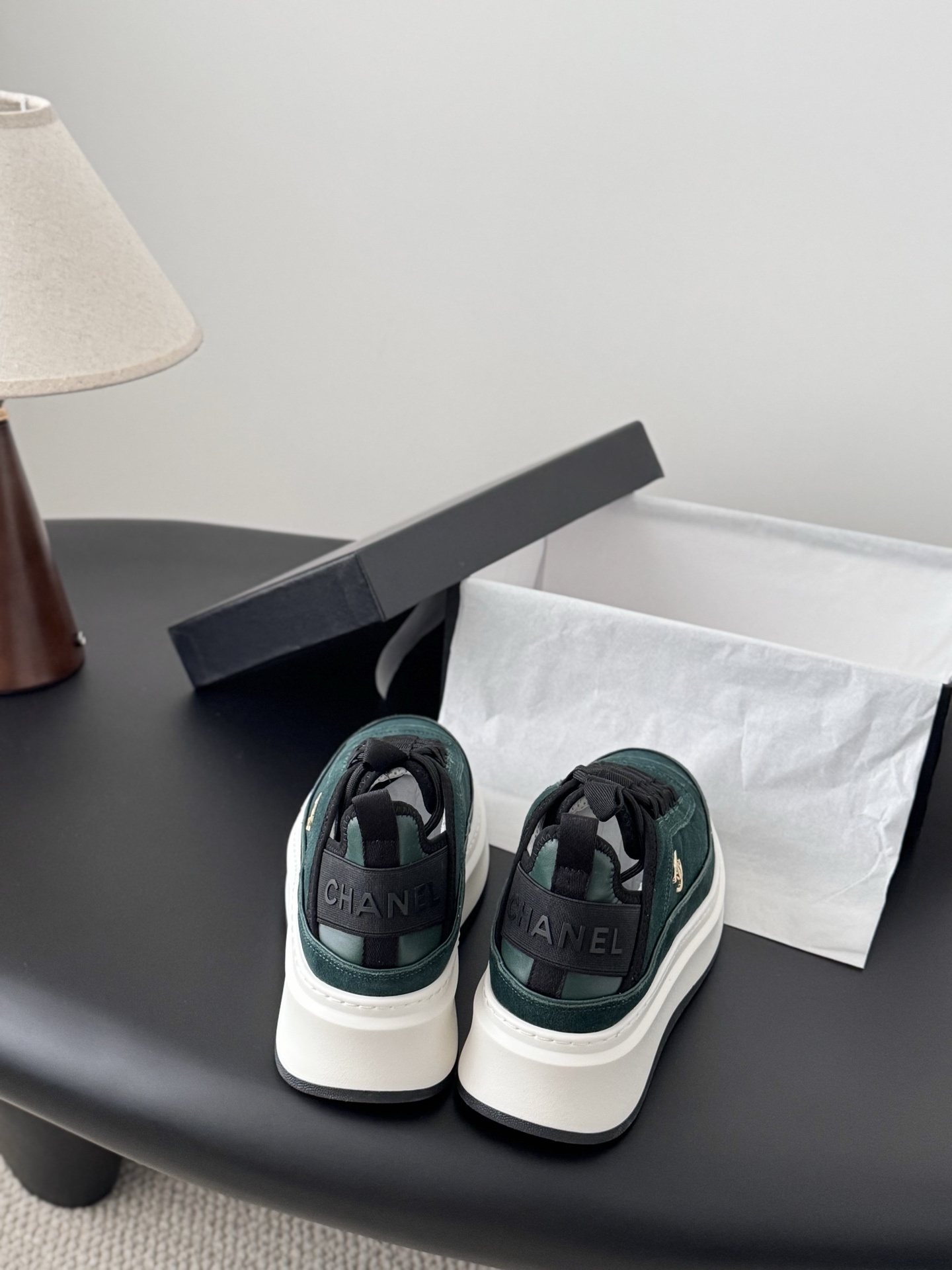 A pair of Chanel dark green suede sneakers with white platform soles and gold CC logos placed next to a black Chanel shoebox on a table. - Hình 5