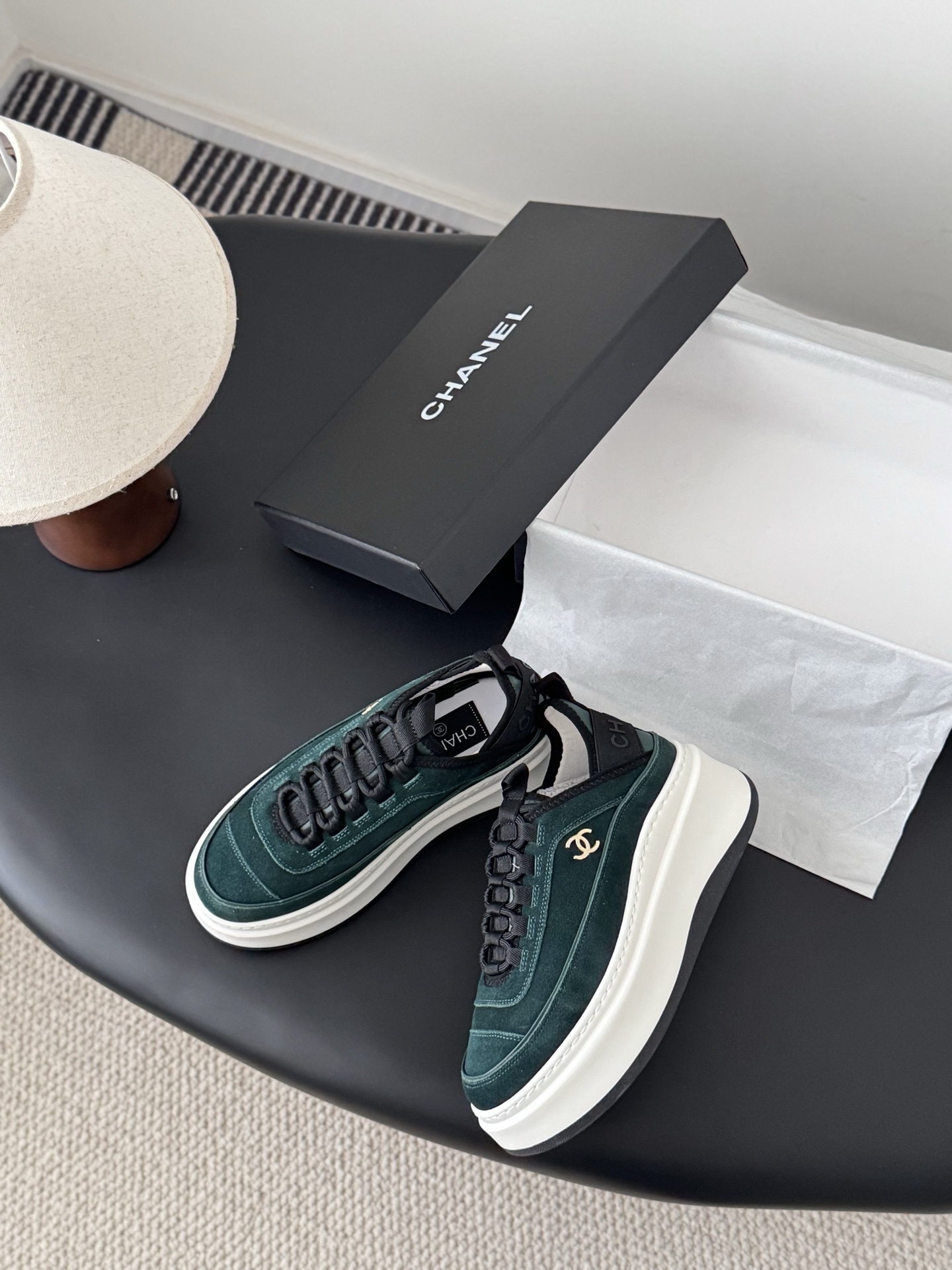 A pair of Chanel dark green suede sneakers with white platform soles and gold CC logos placed next to a black Chanel shoebox on a table. - Hình 3