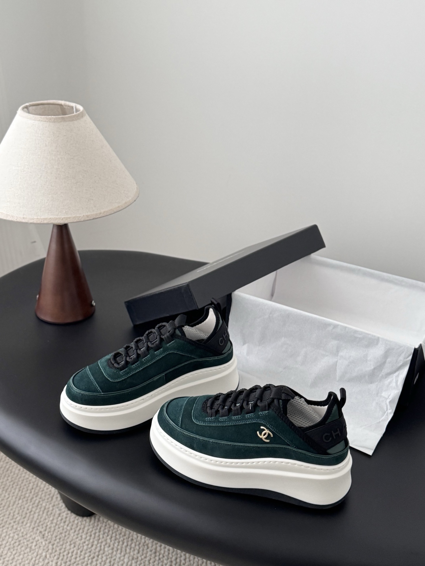 A pair of Chanel dark green suede sneakers with white platform soles and gold CC logos placed next to a black Chanel shoebox on a table. - Hình 2