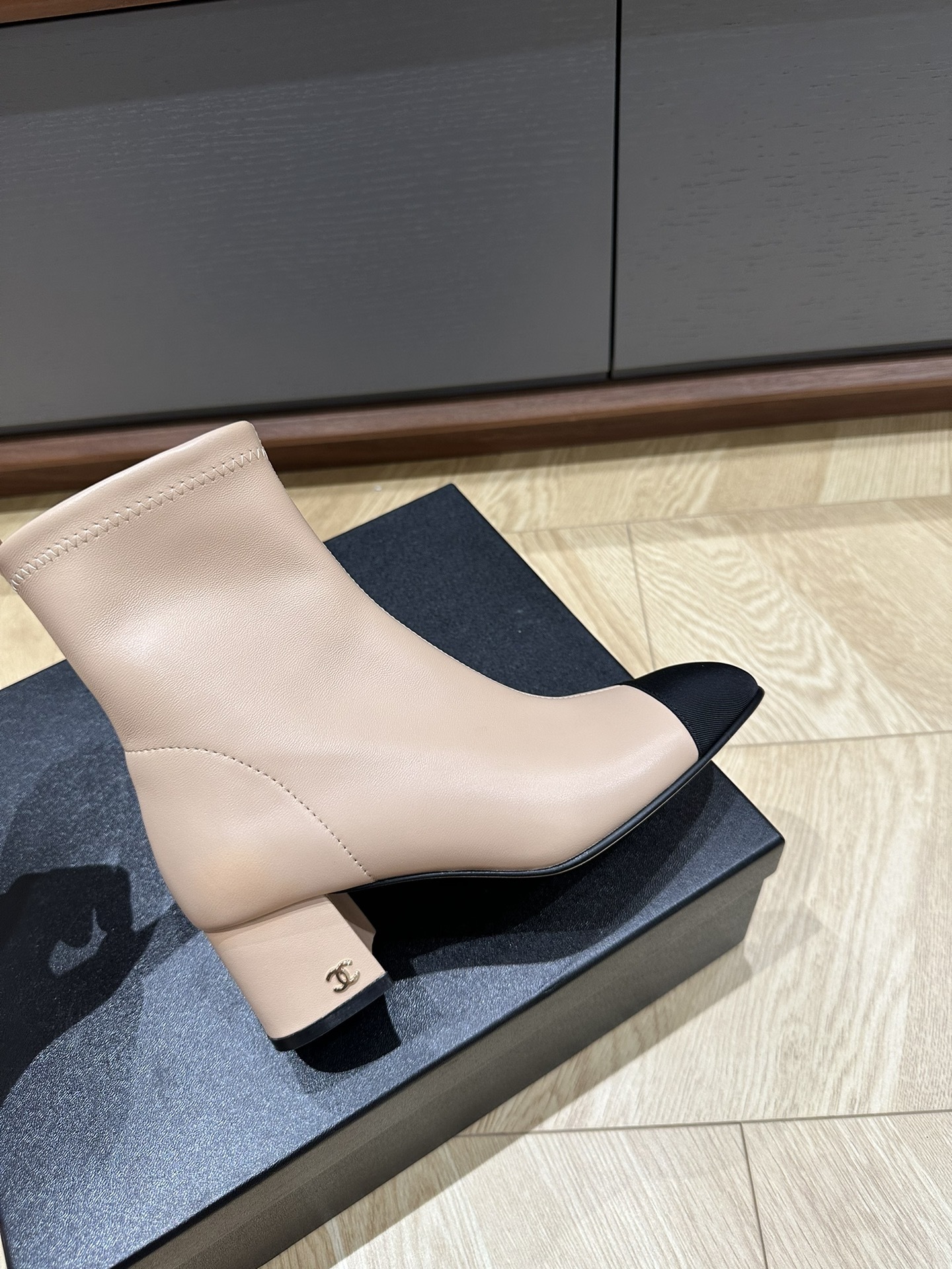 A pair of elegant beige ankle boots with black fabric cap toes and block heels placed on a black shoe box. - Hình 9