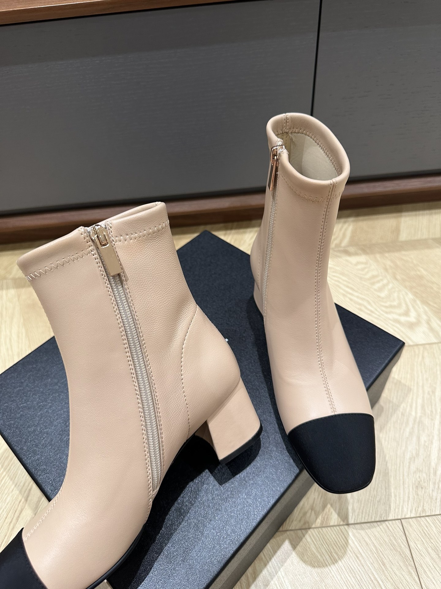 A pair of elegant beige ankle boots with black fabric cap toes and block heels placed on a black shoe box. - Hình 6