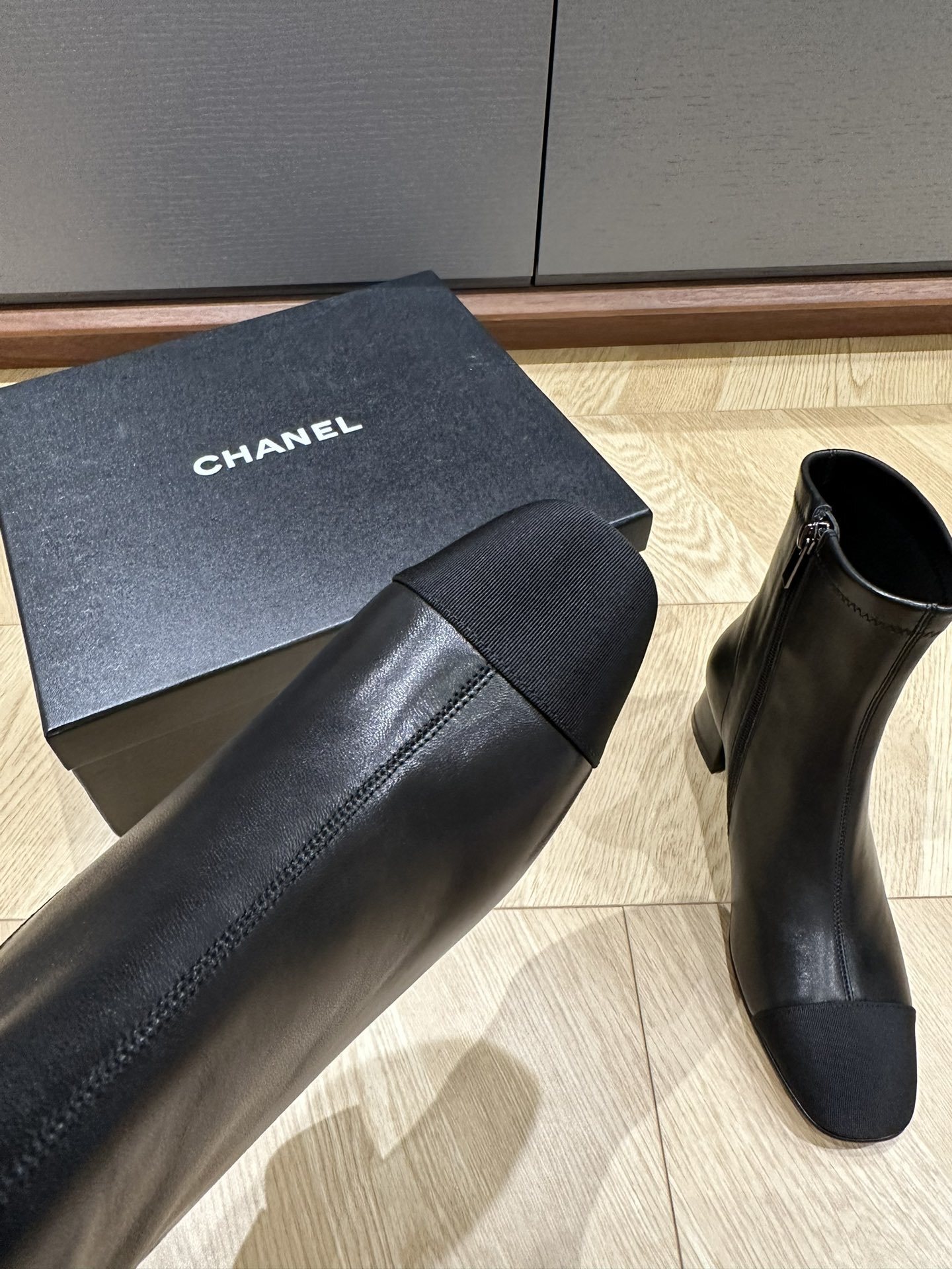 A pair of Chanel black leather ankle boots with grosgrain cap toes and silver CC logo on the block heel, displayed on a Chanel shoe box. - Hình 8