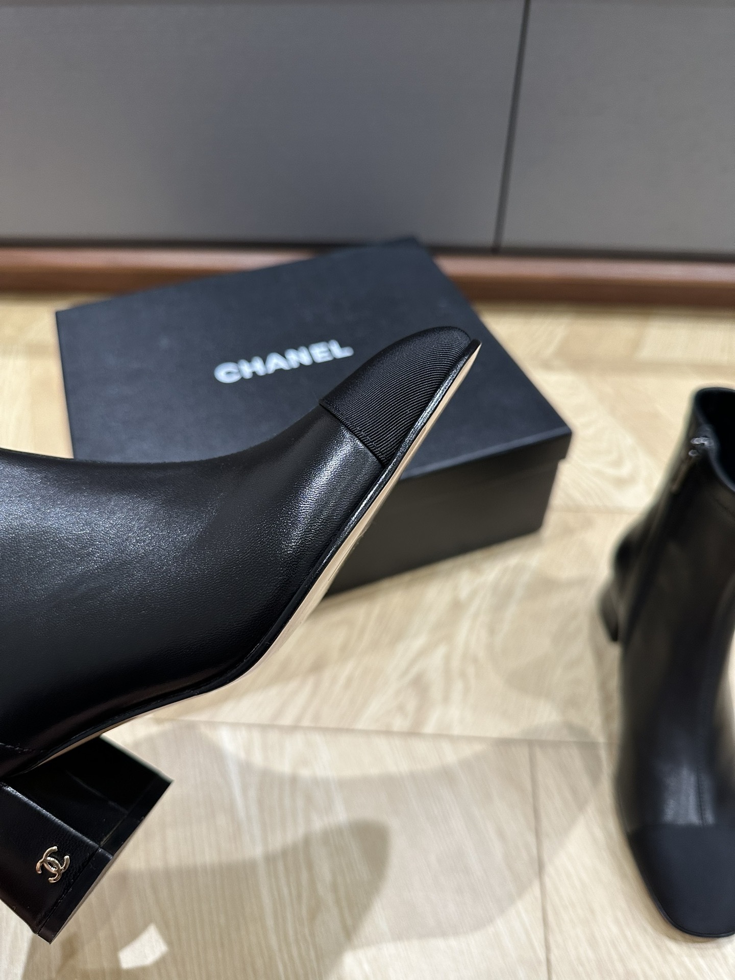 A pair of Chanel black leather ankle boots with grosgrain cap toes and silver CC logo on the block heel, displayed on a Chanel shoe box. - Hình 7
