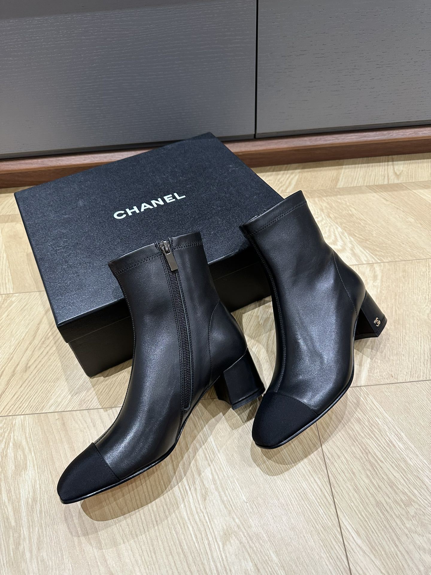 A pair of Chanel black leather ankle boots with grosgrain cap toes and silver CC logo on the block heel, displayed on a Chanel shoe box. - Hình 5