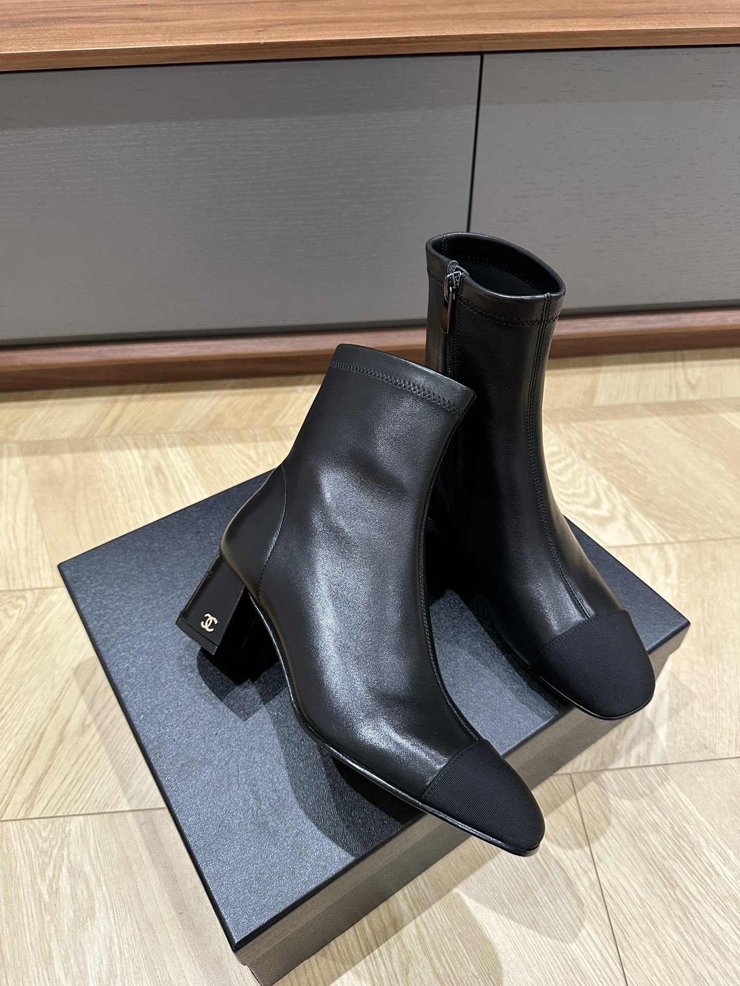 A pair of Chanel black leather ankle boots with grosgrain cap toes and silver CC logo on the block heel, displayed on a Chanel shoe box. - Hình 4