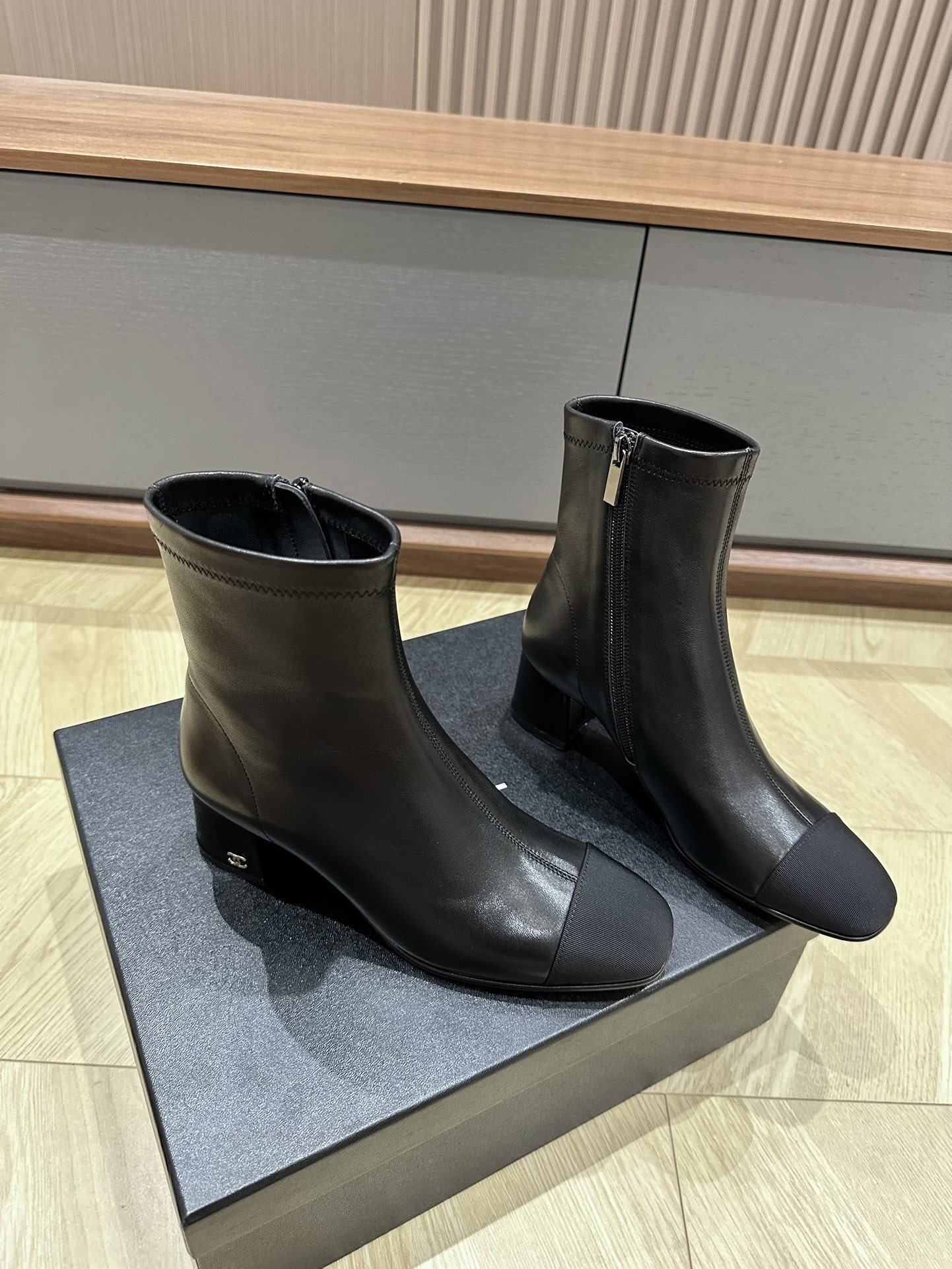 A pair of Chanel black leather ankle boots with grosgrain cap toes and silver CC logo on the block heel, displayed on a Chanel shoe box. - Hình 3