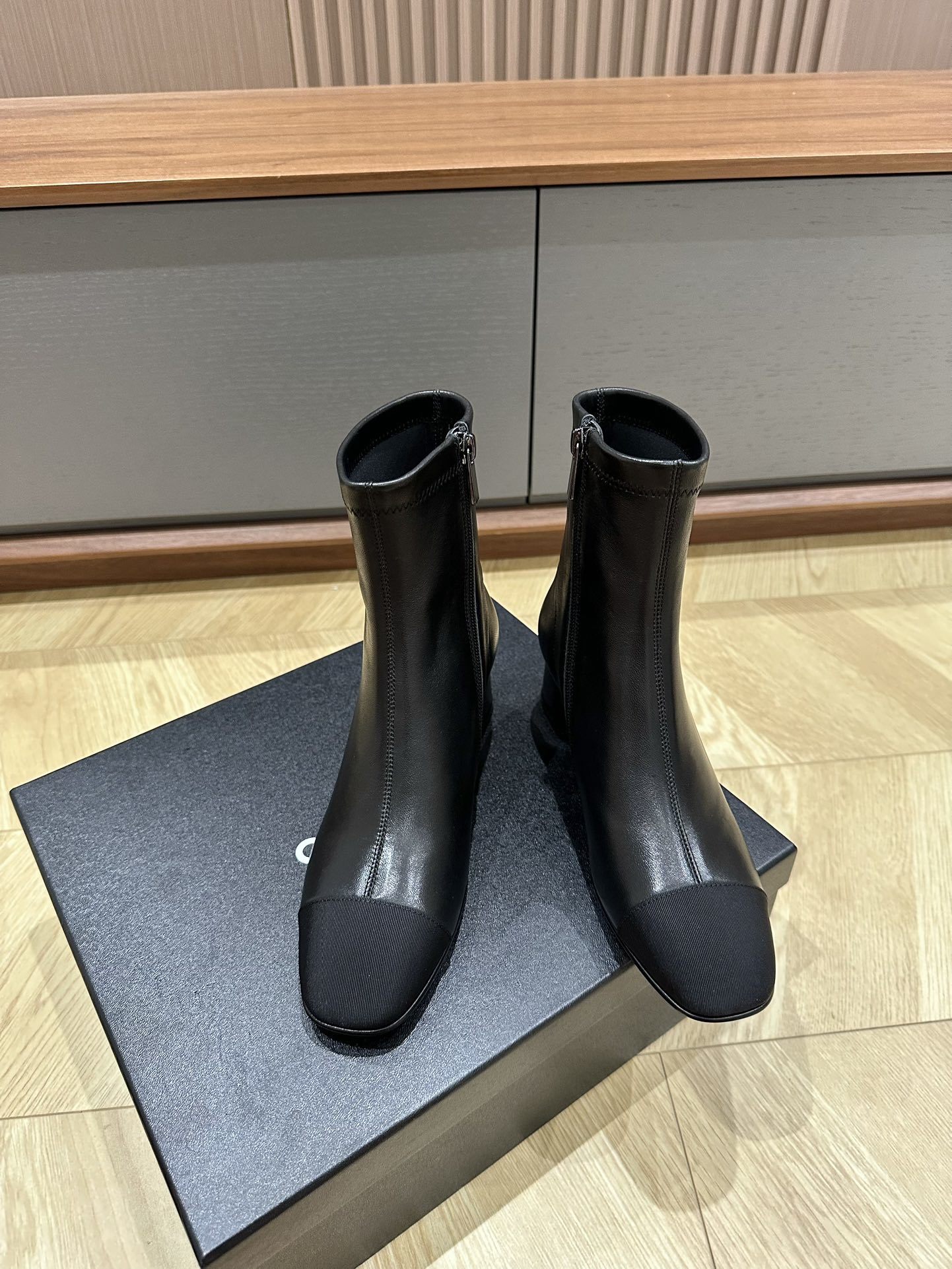 A pair of Chanel black leather ankle boots with grosgrain cap toes and silver CC logo on the block heel, displayed on a Chanel shoe box. - Hình 2