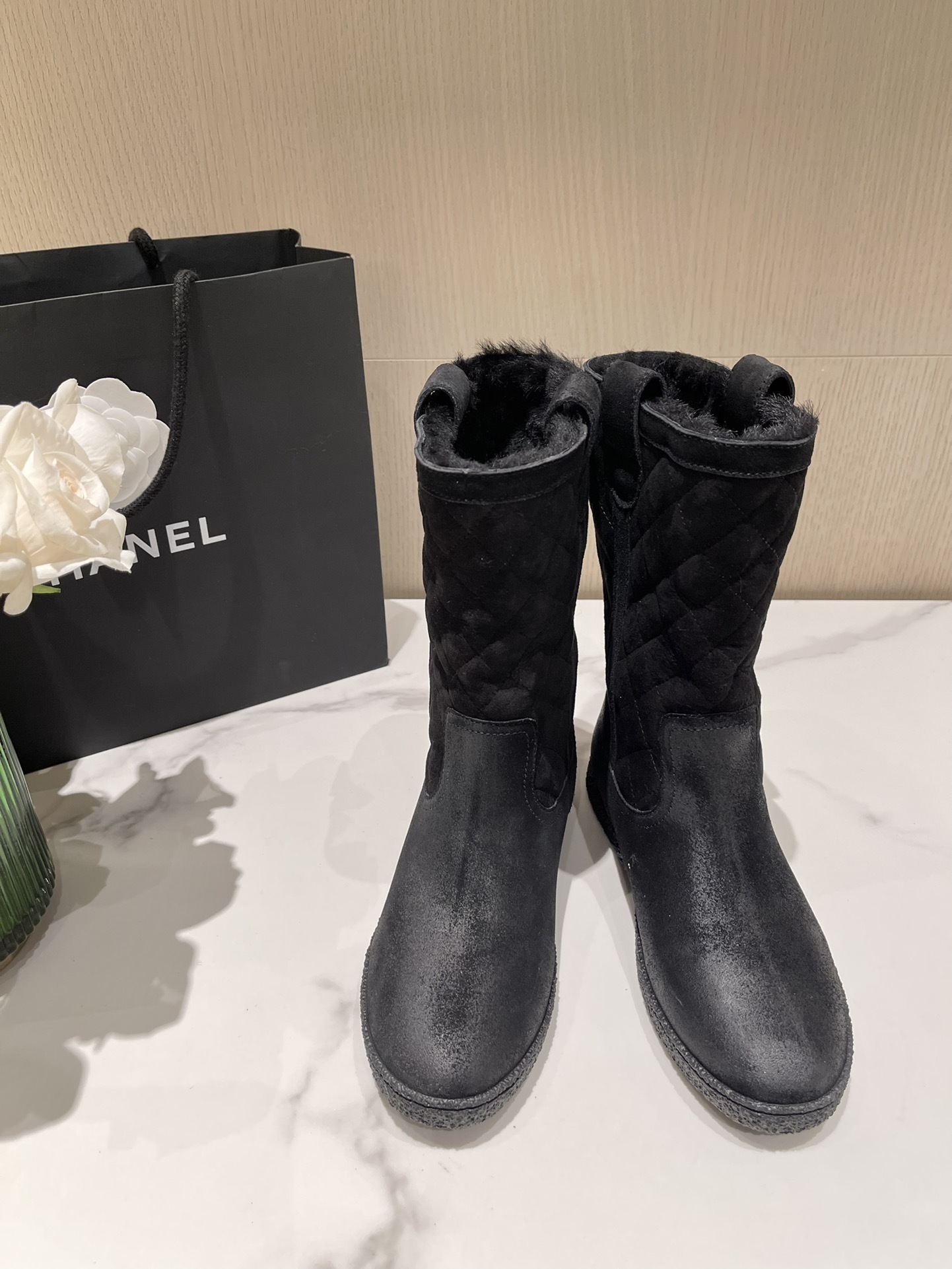 A pair of black Chanel quilted suede winter boots with fur lining and silver CC logo detail on a marble tabletop. - Hình 2