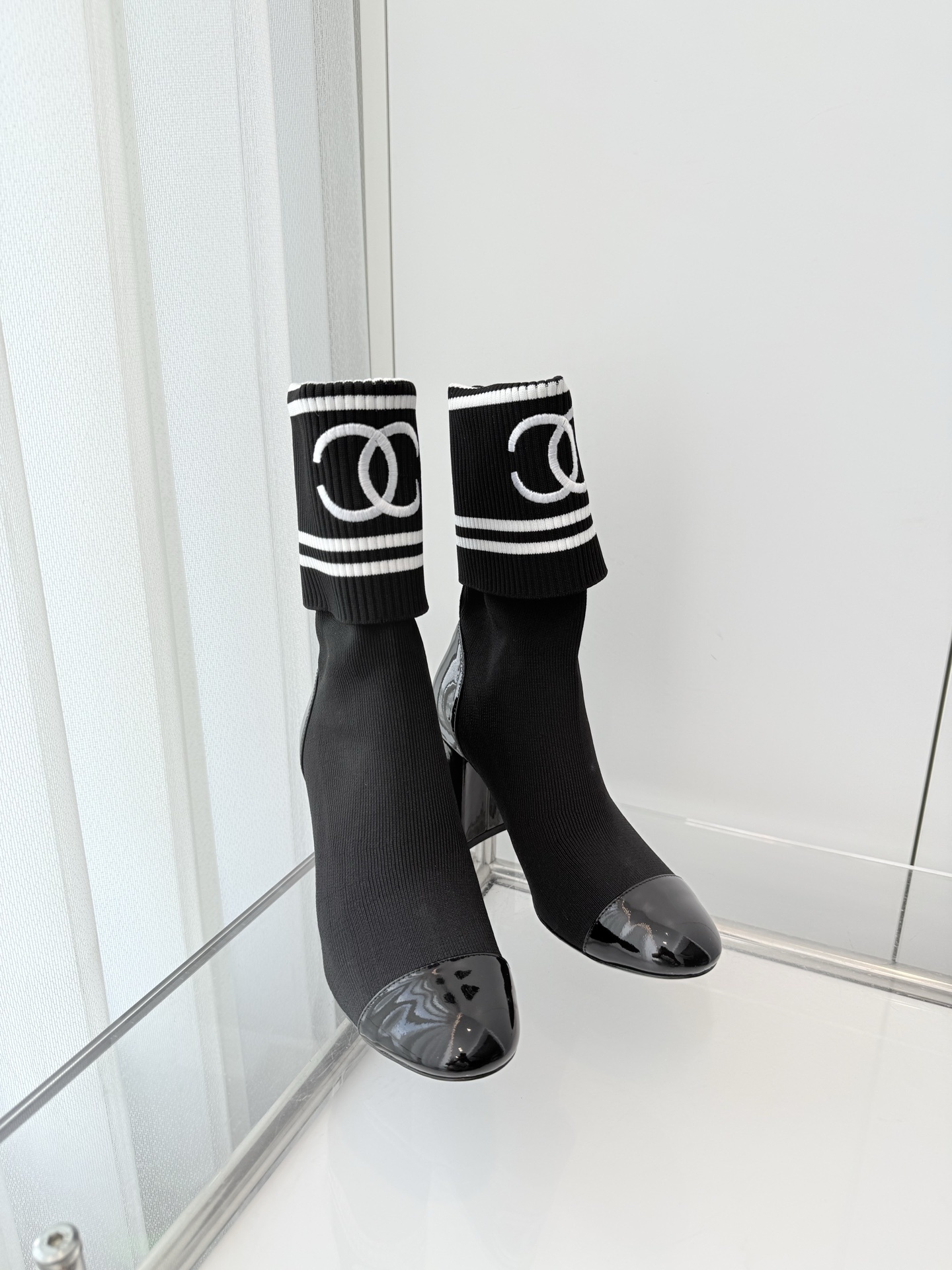 A pair of elegant black knit sock boots with patent leather cap toes and white logo fold-over cuffs displayed on a glass shelf. - Hình 8
